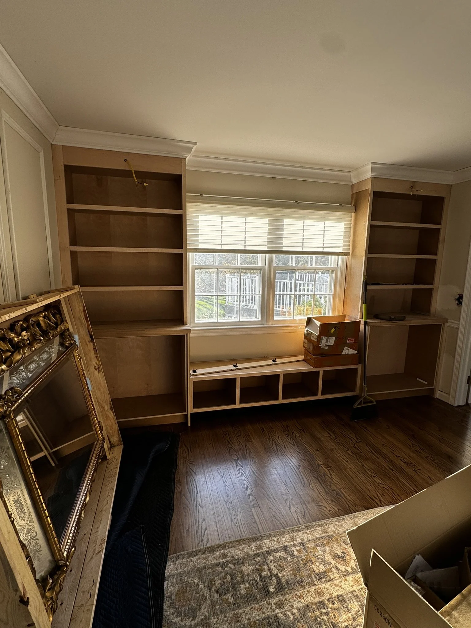 Empty wooden bookshelves with a box and cleaning tools in front of a window in a room under renovation.
