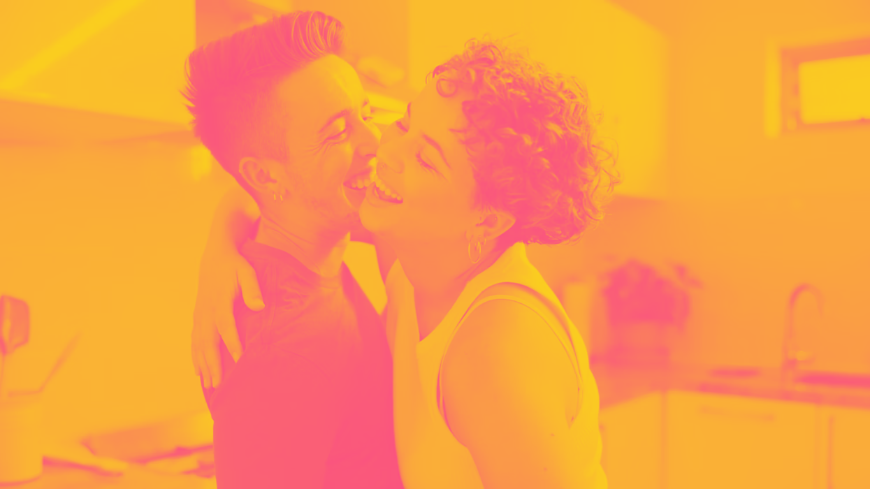 Two masculine transgender people hugging and smiling, in a kitchen setting with warm lighting.