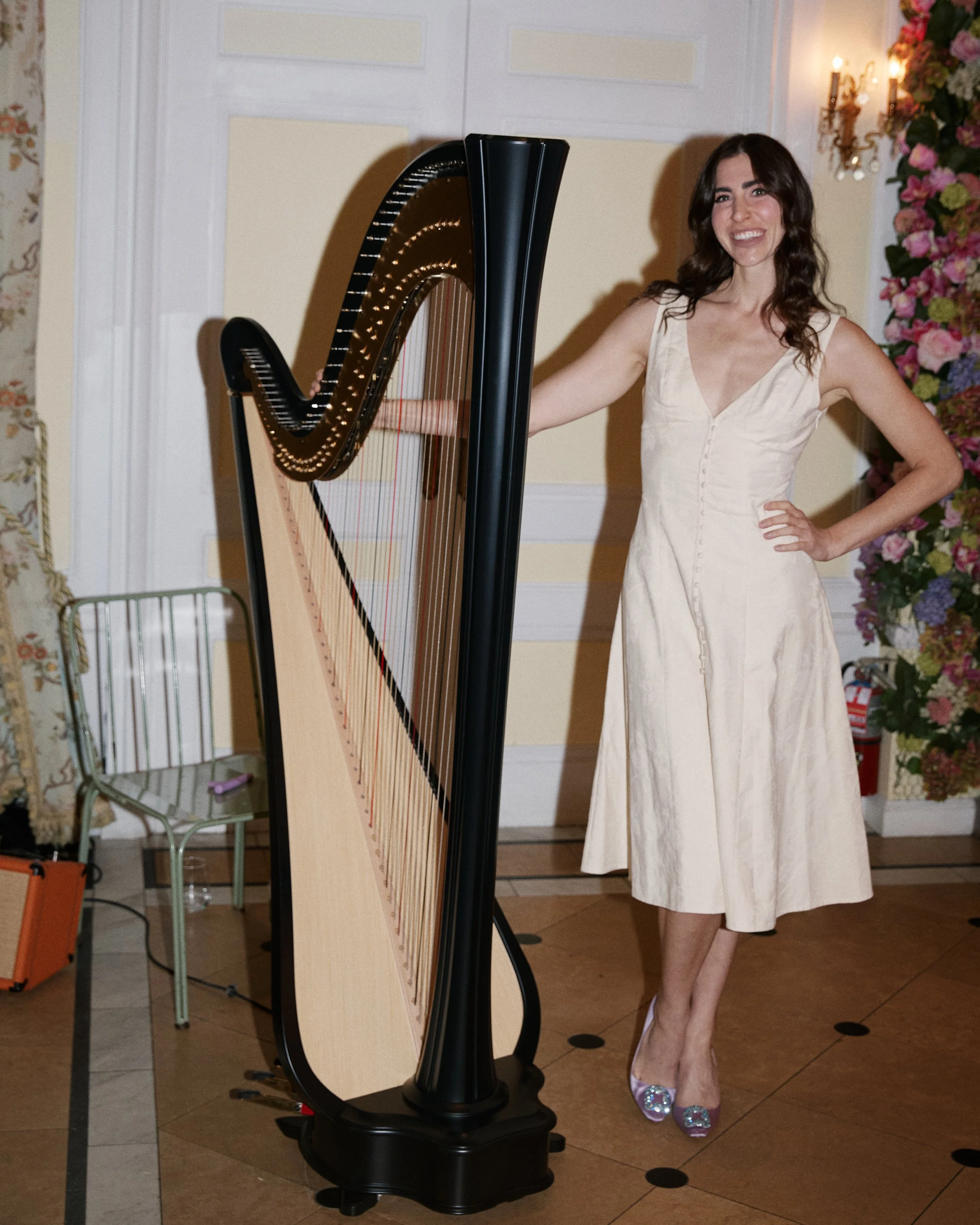 Woman in a cream dress standing next to a large harp in a decorated room with flowers and a chandelier.