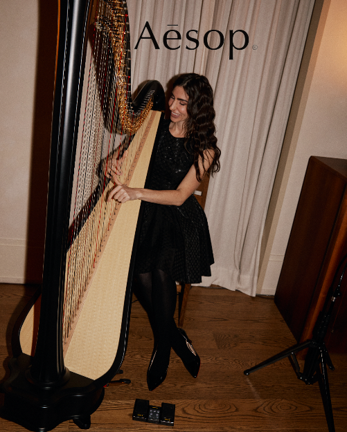 A woman with long wavy dark hair wearing a black dress and black tights playing a large wooden harp indoors in a room with curtains and wooden flooring.