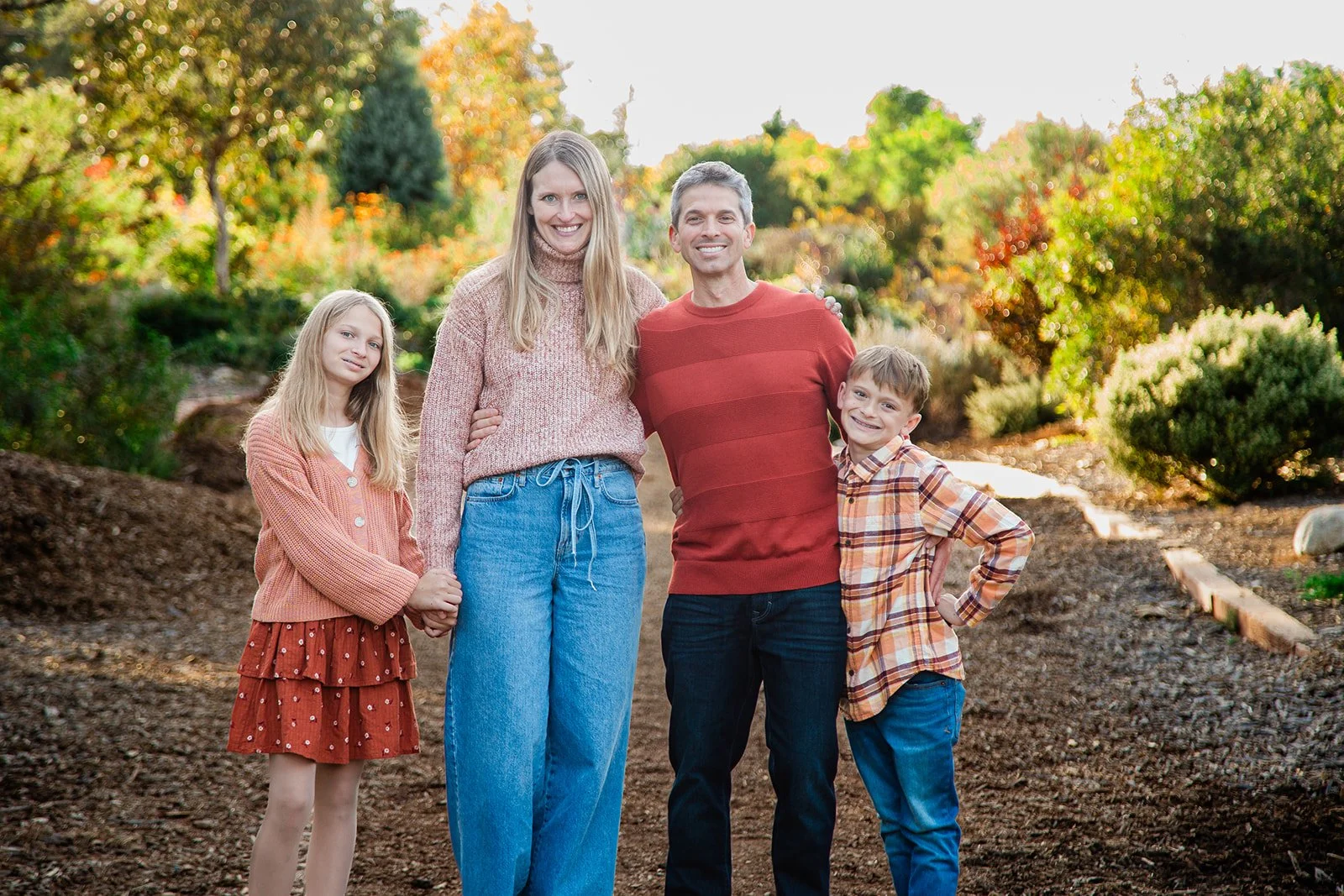 A family is posing for photos at the Urban Forest in Huntington Beach, CA during the fall months with sweaters and fall colored clothing on, and green trees and fall colored trees behind them