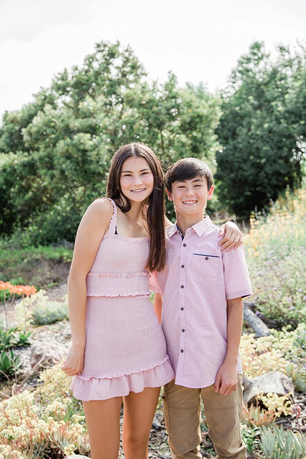Siblings taking photos in the Urban Forest park in Huntington Beach, CA during the superbloom with beautiful green trees and flowering bushes behind them while they're hugging and smiling together.