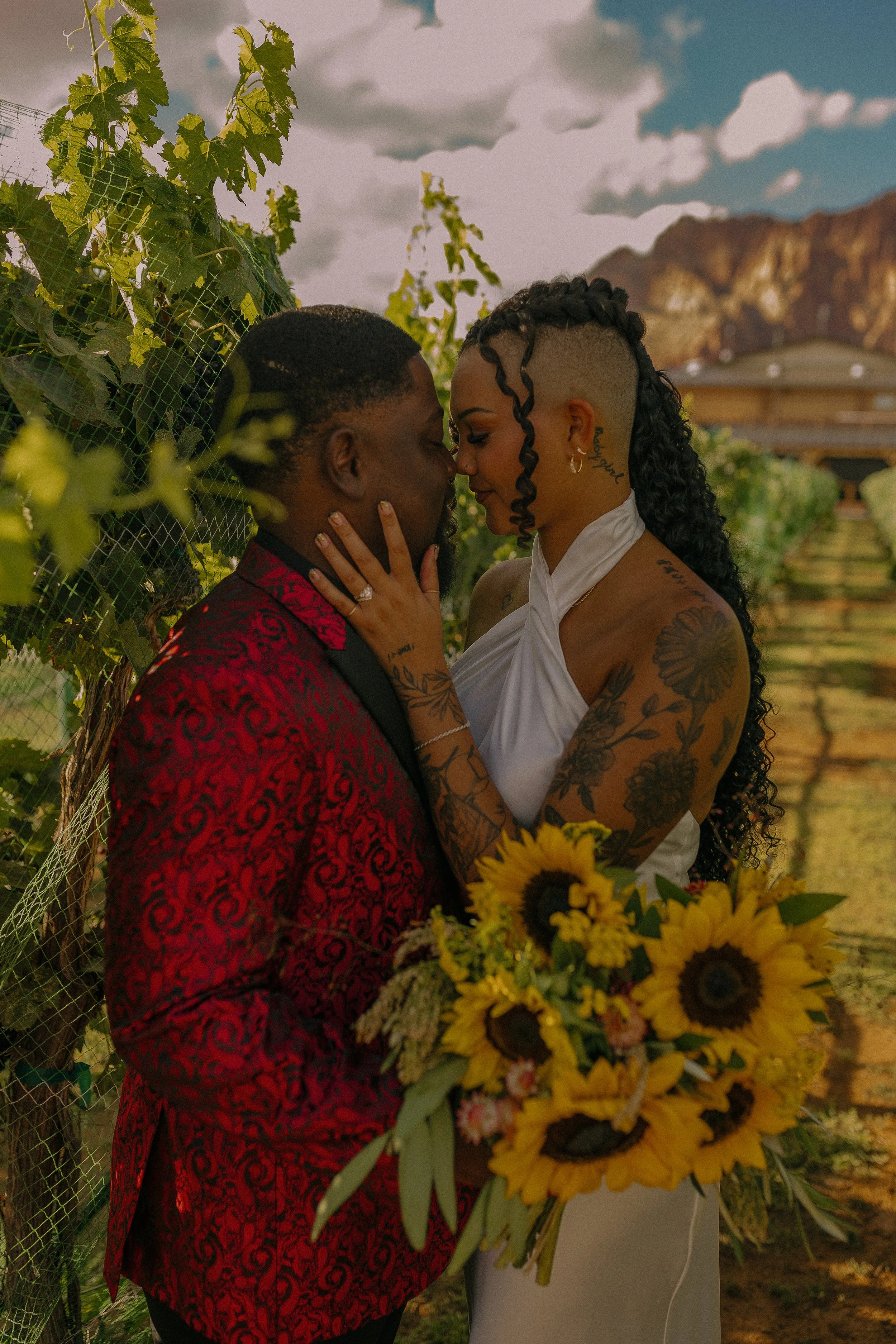 A couple embracing in a vineyard, the woman holds a bouquet of sunflowers.