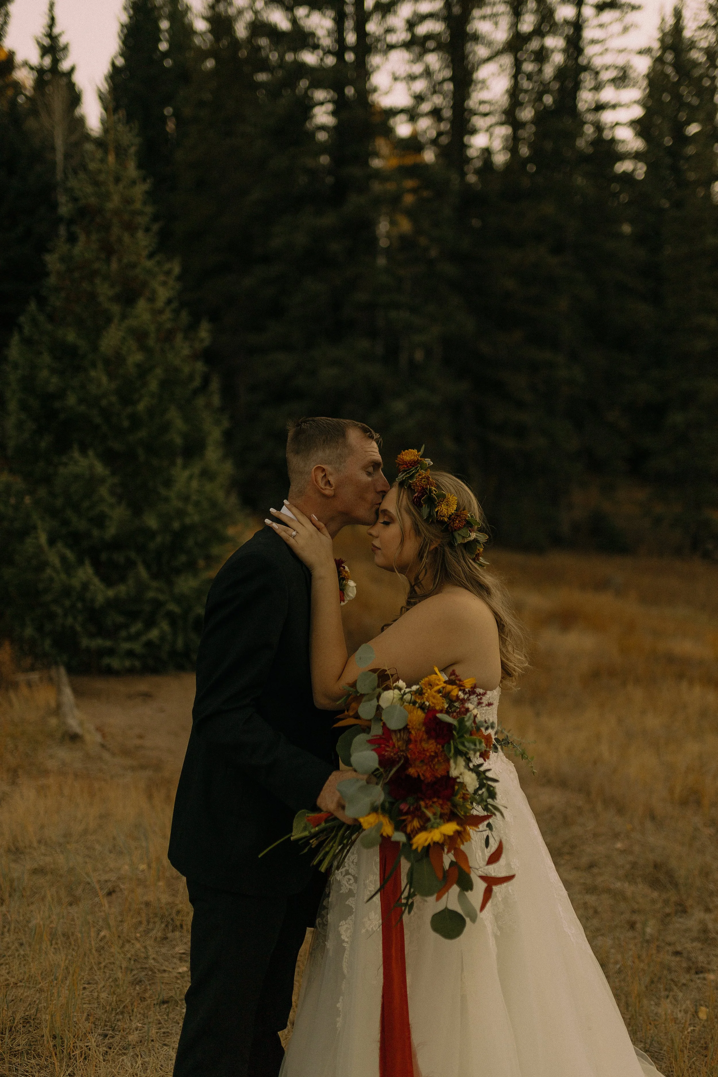 A bride and groom embrace in an outdoor forest setting wearing a wedding dress and suit. The bride holds a bouquet of flowers and wears a floral crown.