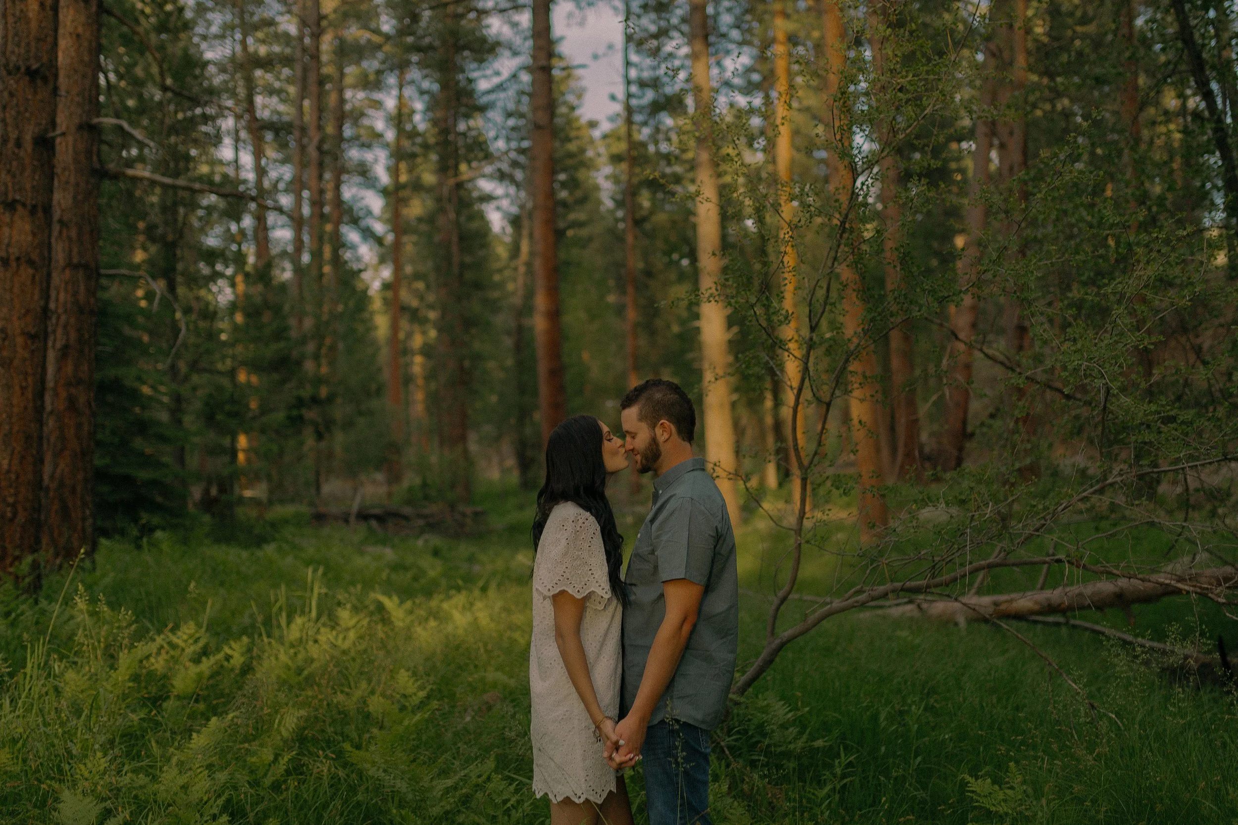Couple kissing in a forest surrounded by trees and greenery.
