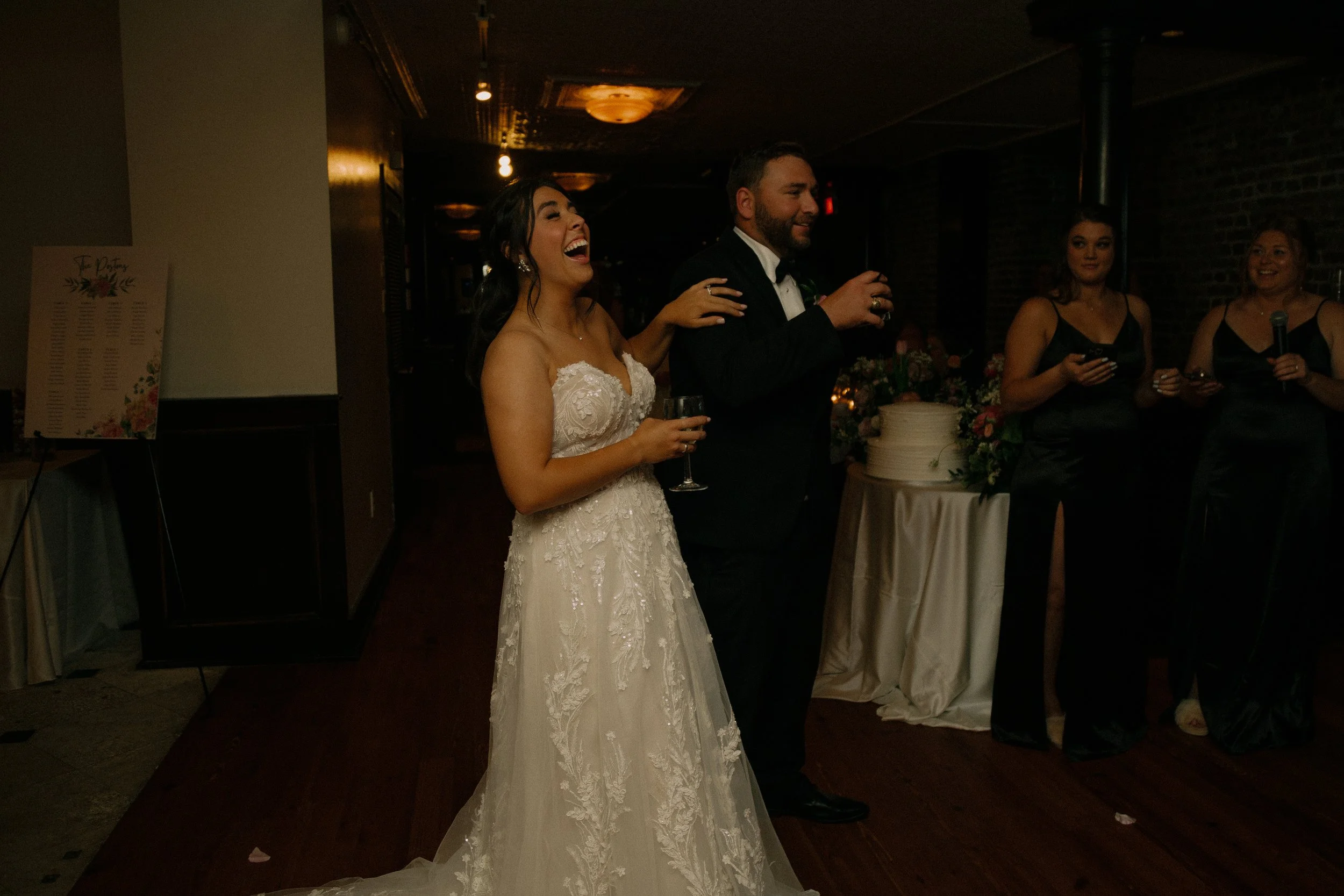 Bride and groom at wedding reception, holding glasses, standing near a table with a cake, with two bridesmaids nearby in black dresses.