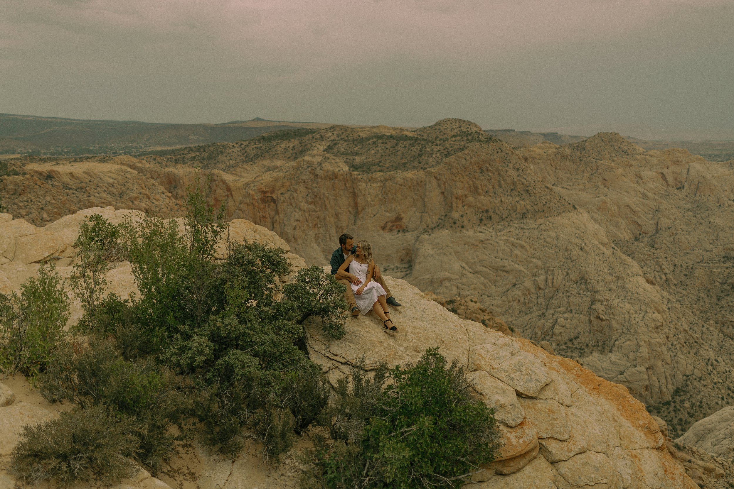 Couple sitting on a rocky cliff with green shrubs, overlooking a rugged mountain landscape.