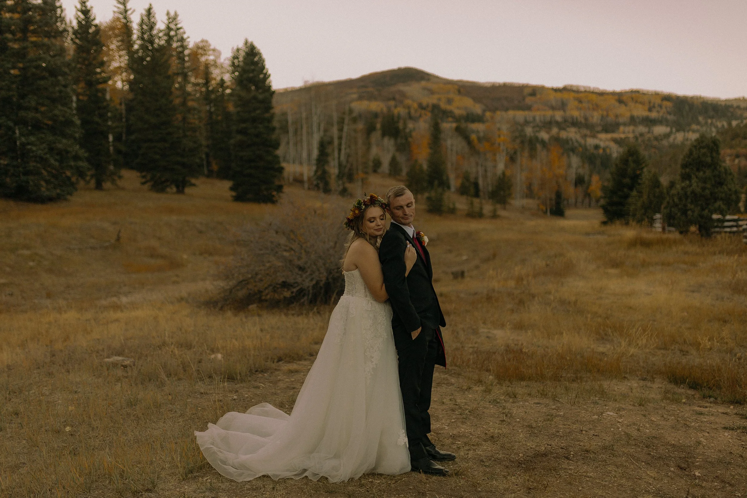 A bride and groom standing in a grassy field with forested hills in the background, the bride wearing a floral crown and a white gown, and the groom in a black suit.