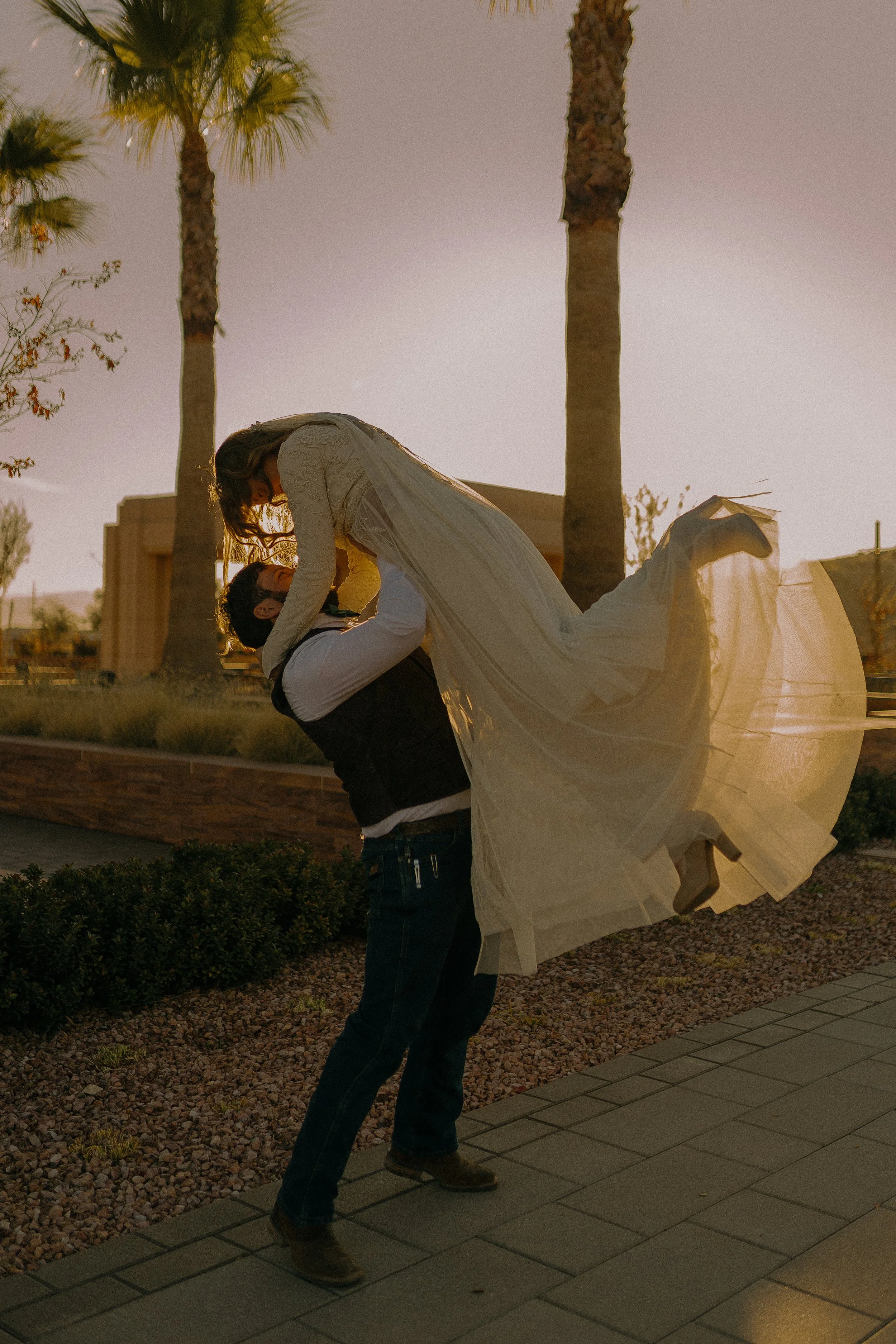 Man lifting woman in white dress outdoors, surrounded by palm trees, during sunset.