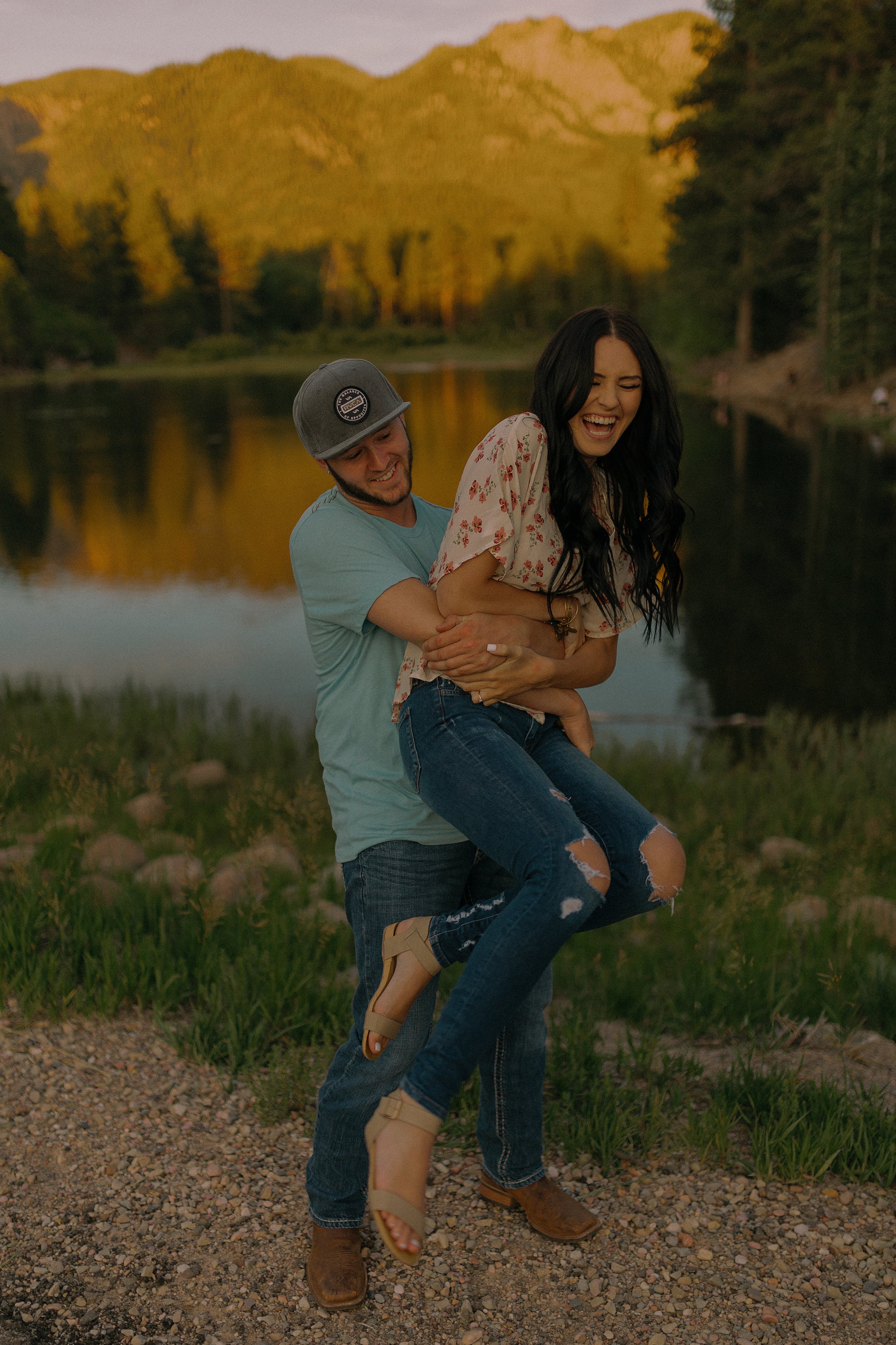 Man playfully lifting a woman near a serene lakeside with mountains in the background.