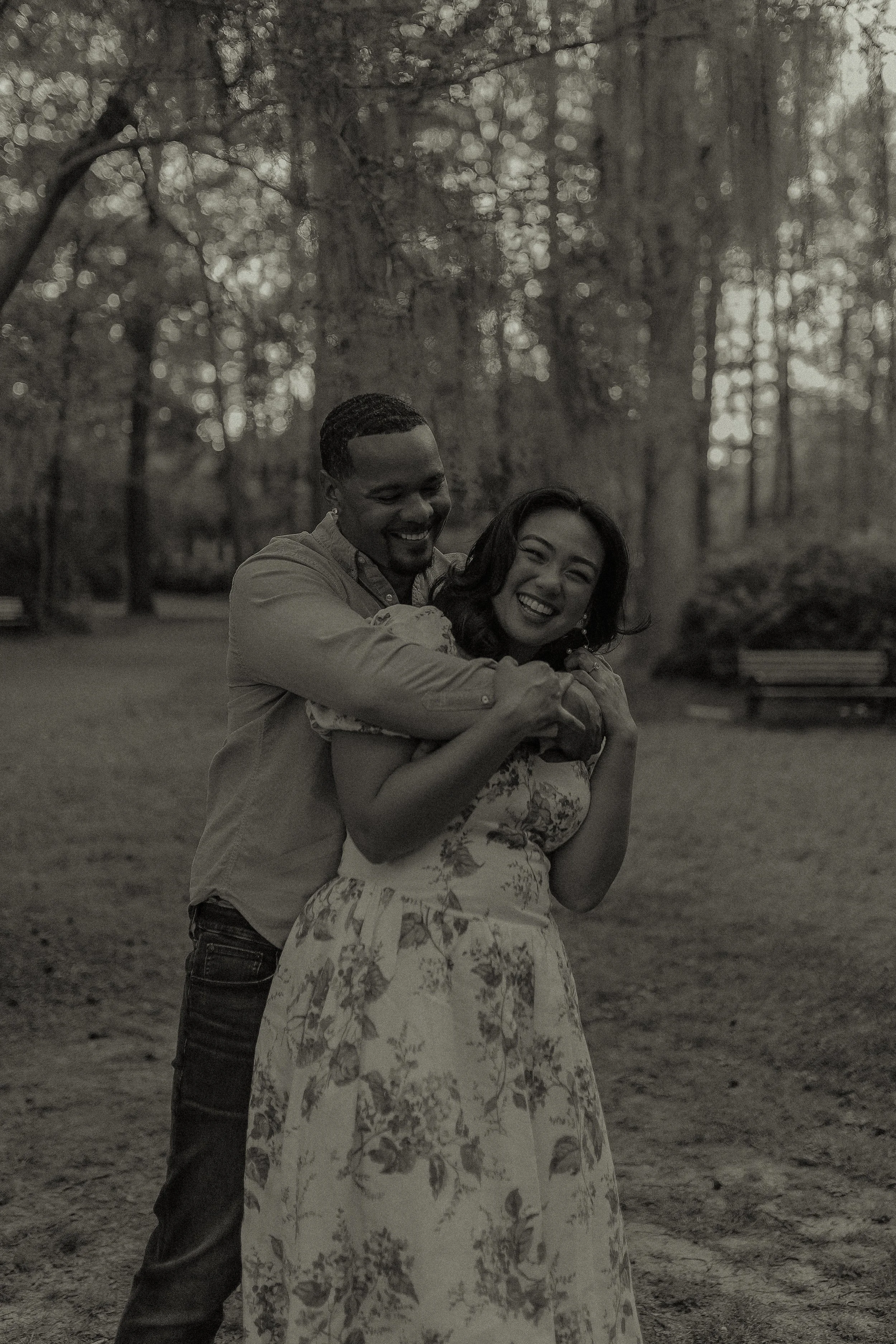 A smiling couple embracing in a wooded park setting, with the woman wearing a floral dress.