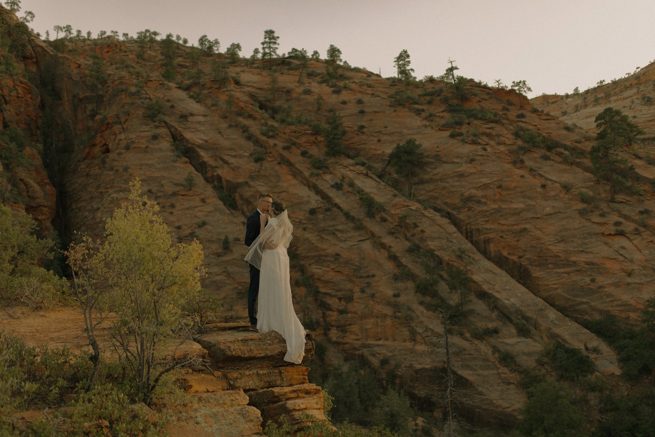 Couple in wedding attire kissing on a cliff edge with rocky landscape and trees in the background.