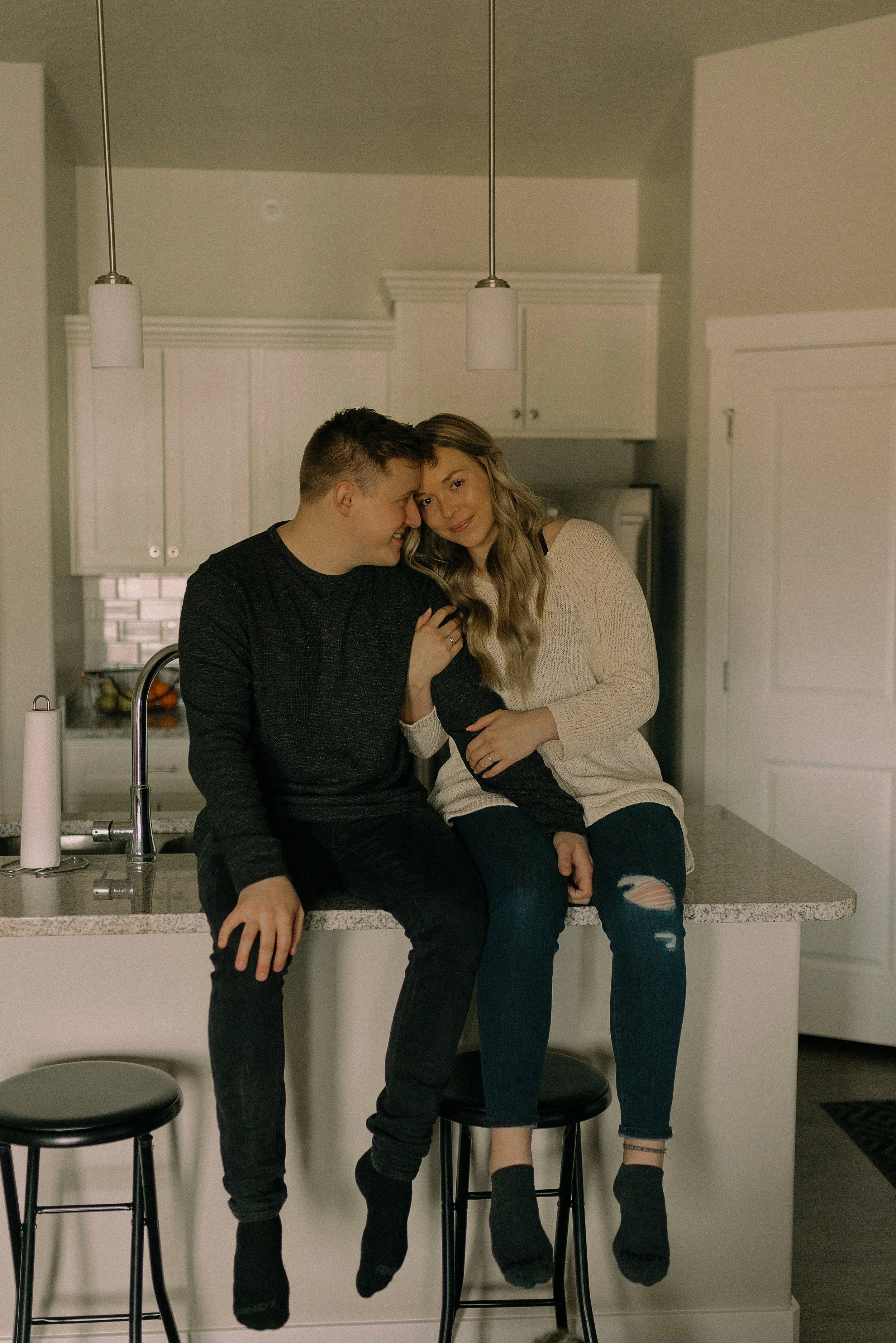 A couple sitting on a kitchen counter, leaning towards each other, in a cozy and affectionate pose. They are indoors in a modern kitchen setting with overhead lights and cabinetry in the background.