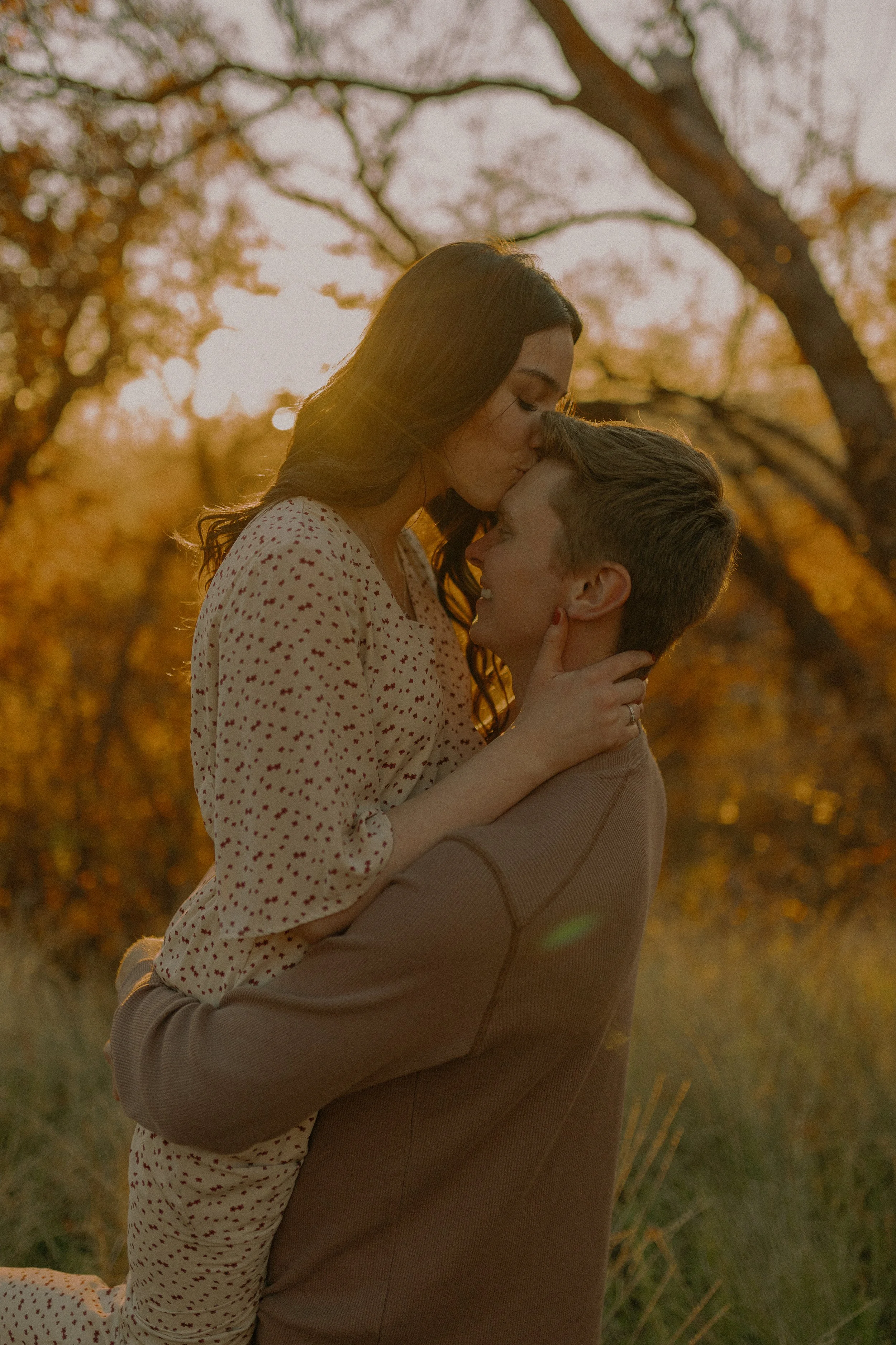 A couple embracing in a field during sunset, with the woman kissing the man's forehead.