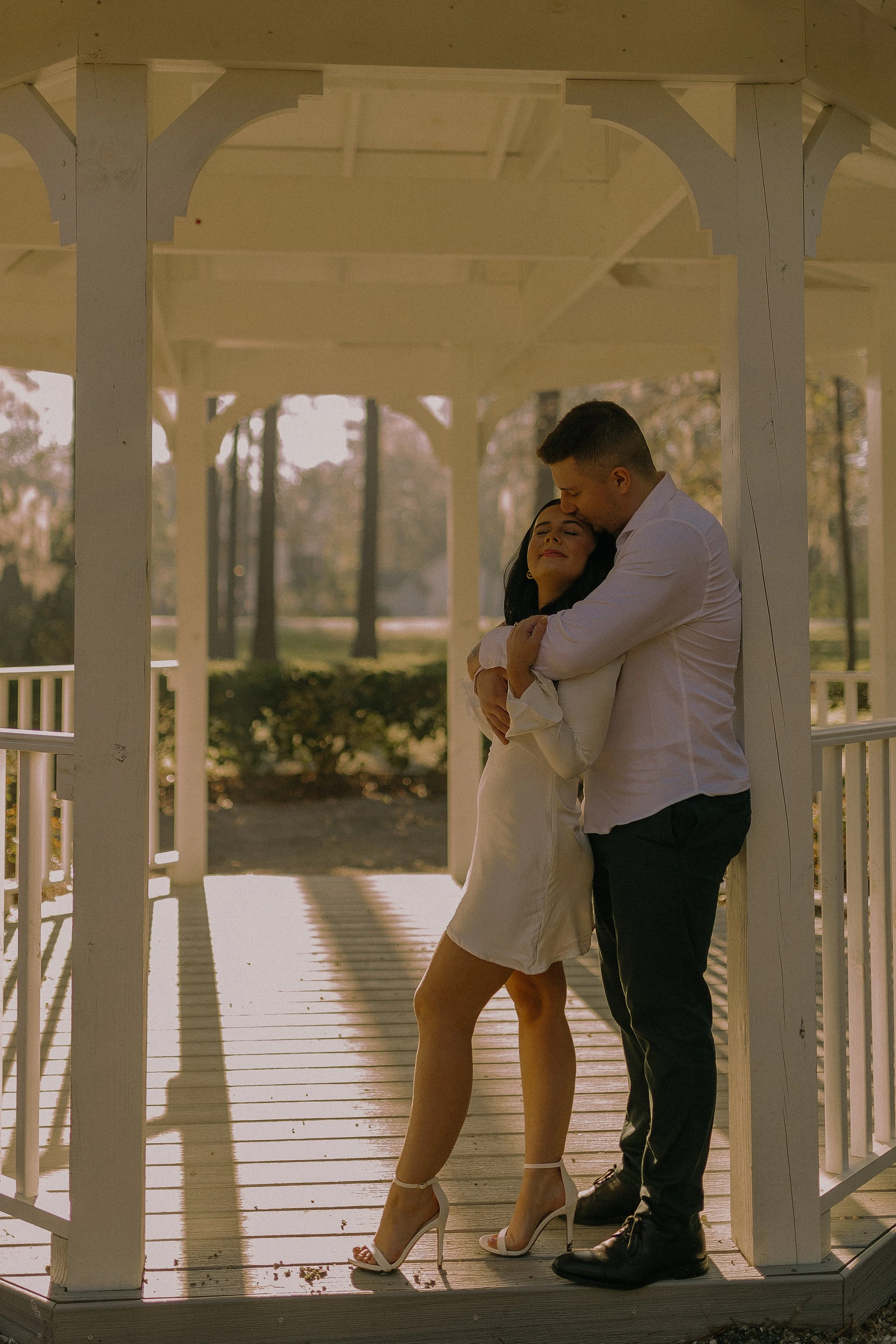 A couple embraces romantically under a gazebo in a sunlit park. The woman is wearing a white dress and heels, and the man is wearing a white shirt and dark pants.