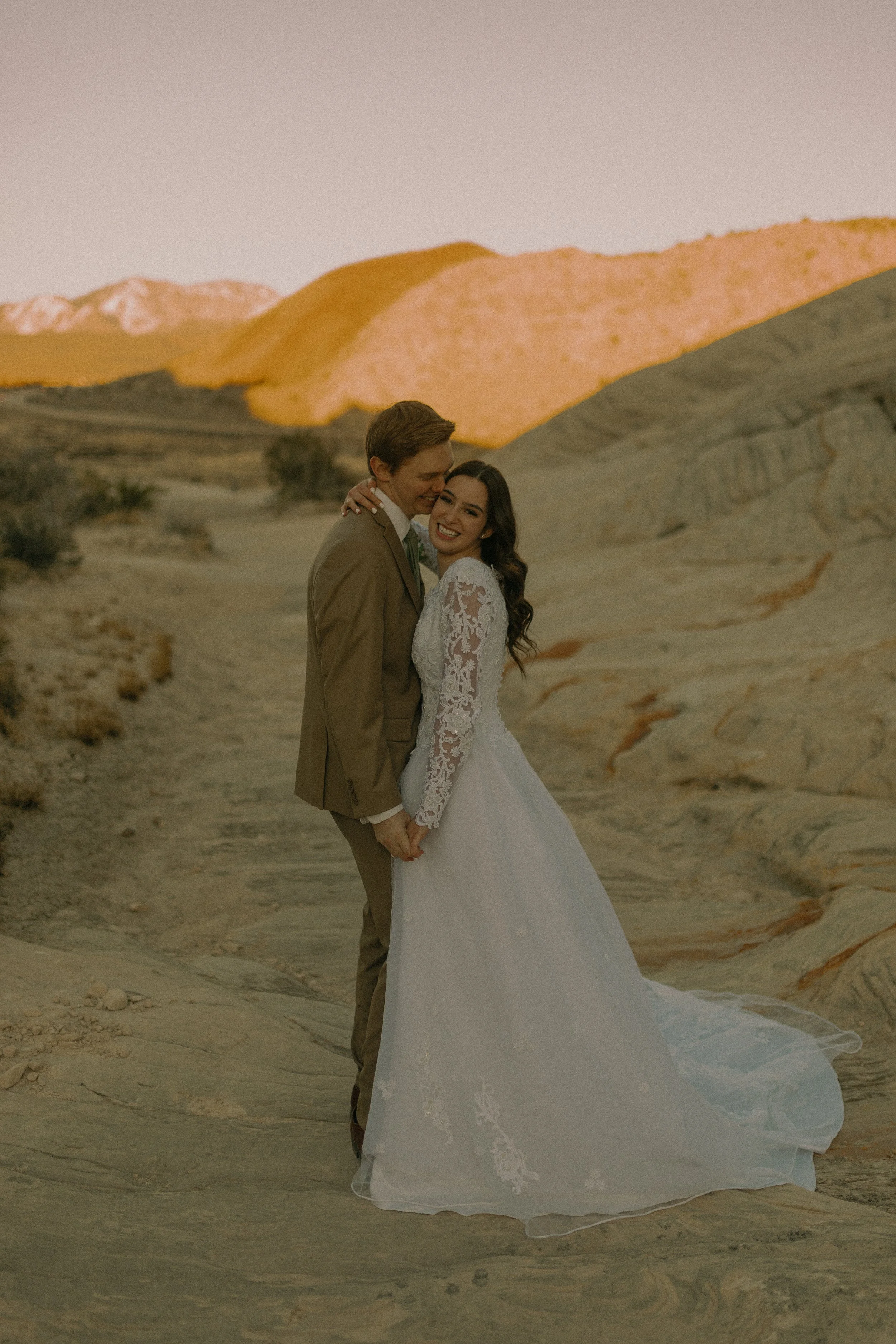 A couple embracing in wedding attire, standing on a rocky landscape with mountains in the background.