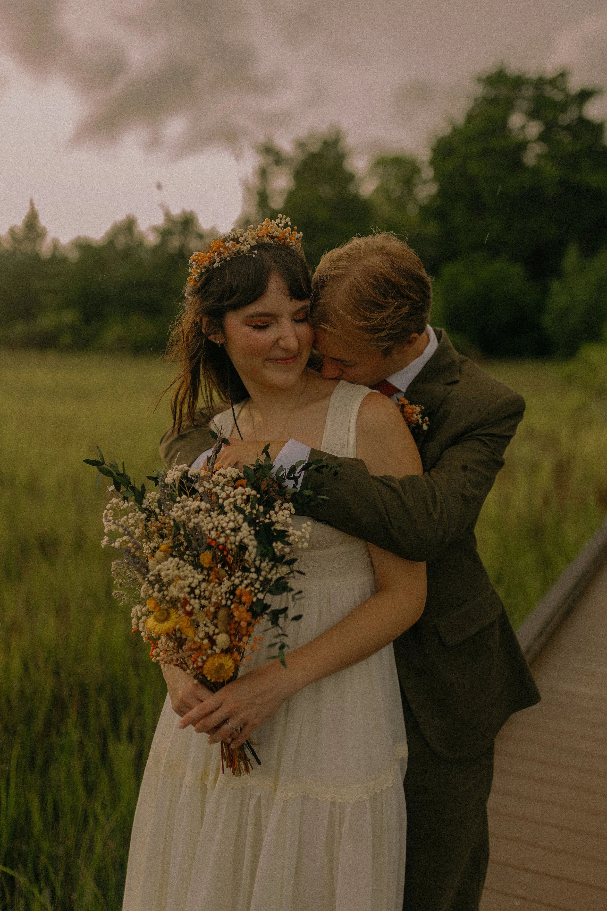 Bride in white dress holding bouquet embraces groom on wooden path in a field