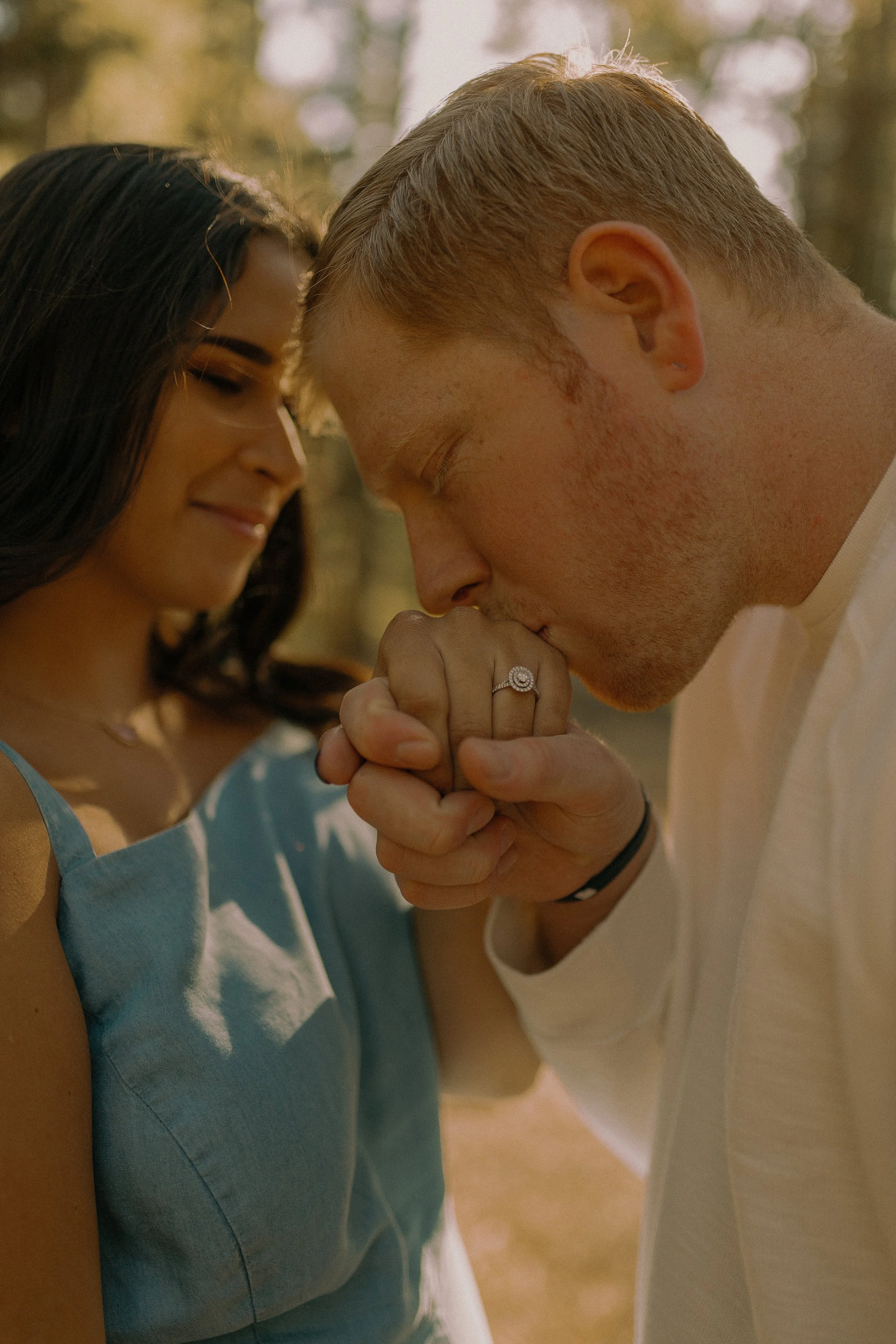 Man kissing woman's hand showing engagement ring in outdoor setting.