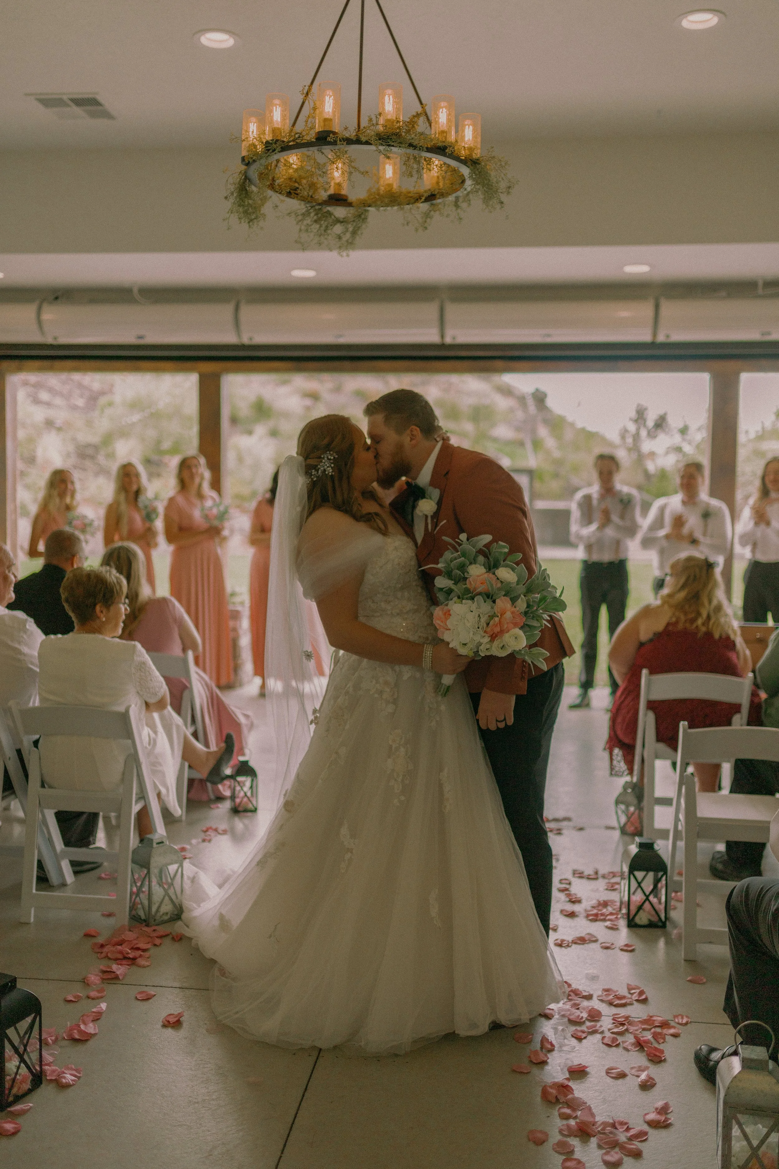 Bride and groom kissing at indoor wedding ceremony, surrounded by guests and floral decorations.