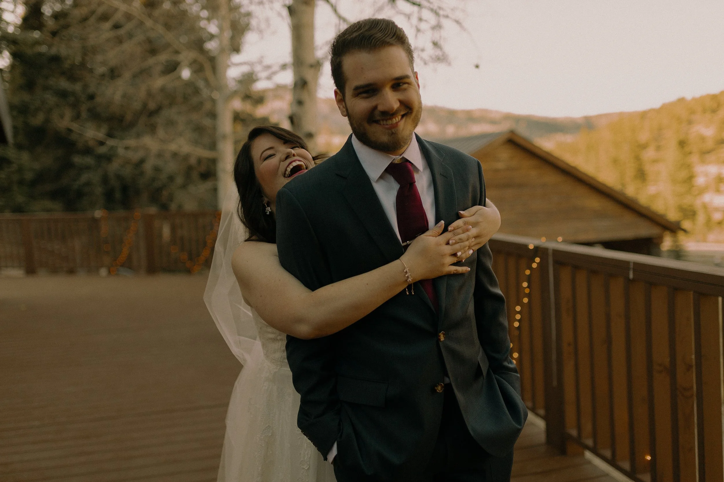Bride playfully hugging groom from behind on a wooden deck, both smiling, with a natural outdoor background.