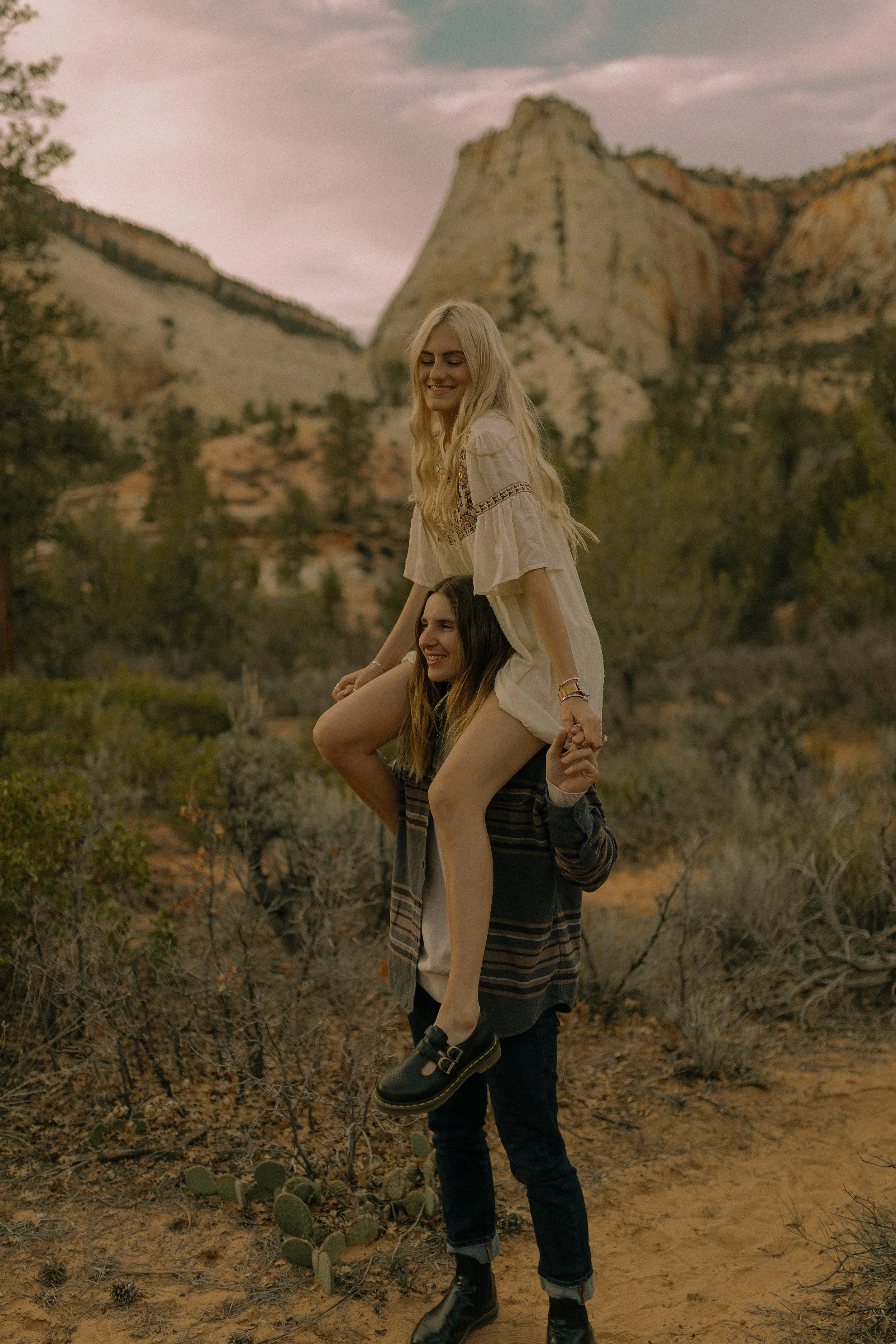 A person with long hair stands outdoors in a mountainous area, carrying another person sitting on their shoulders. The person on top has long blonde hair and is wearing a white dress. The background features rock formations and trees under a cloudy s