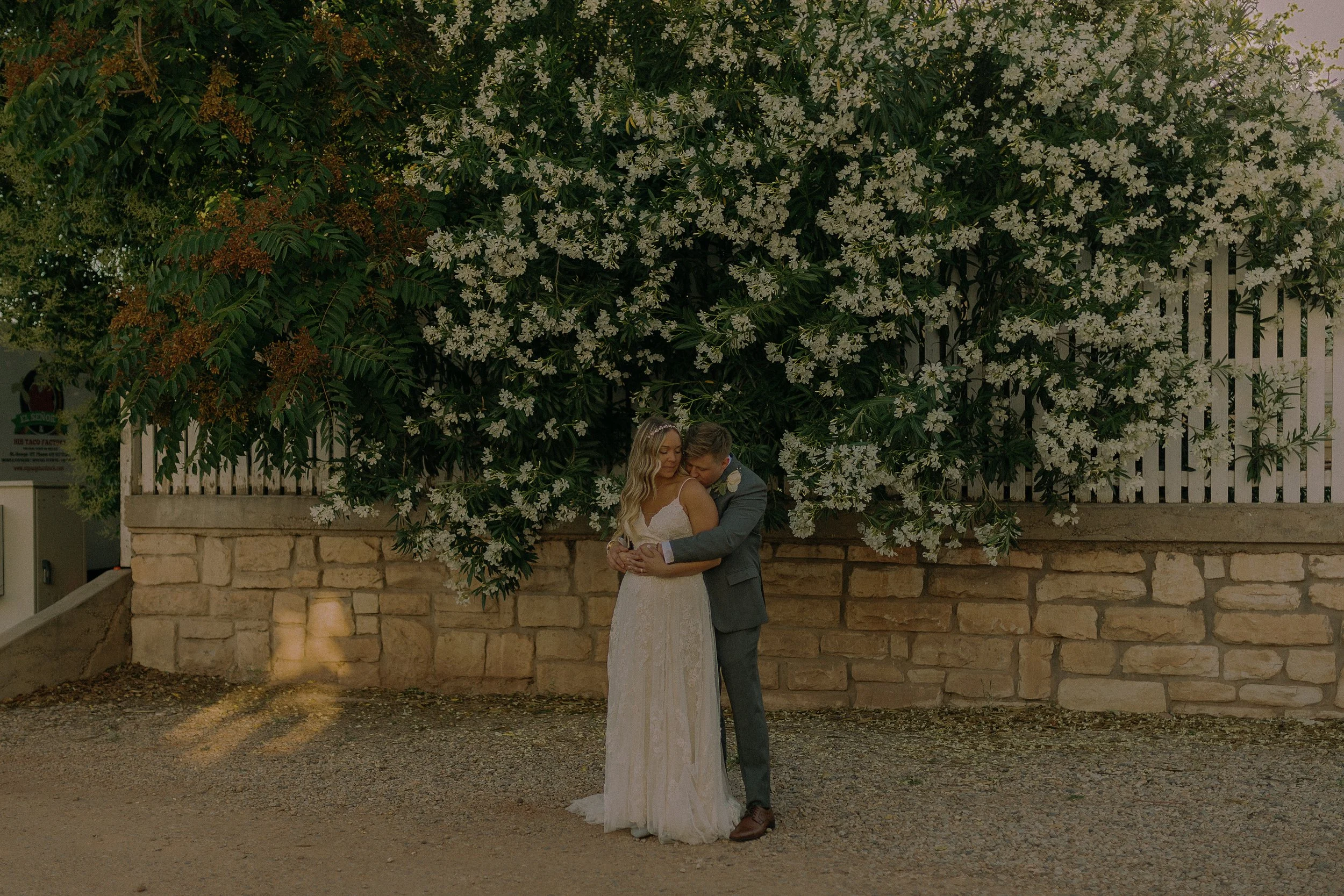 A couple embracing in front of a wall covered with white flowers and greenery, with a stone and picket fence background.
