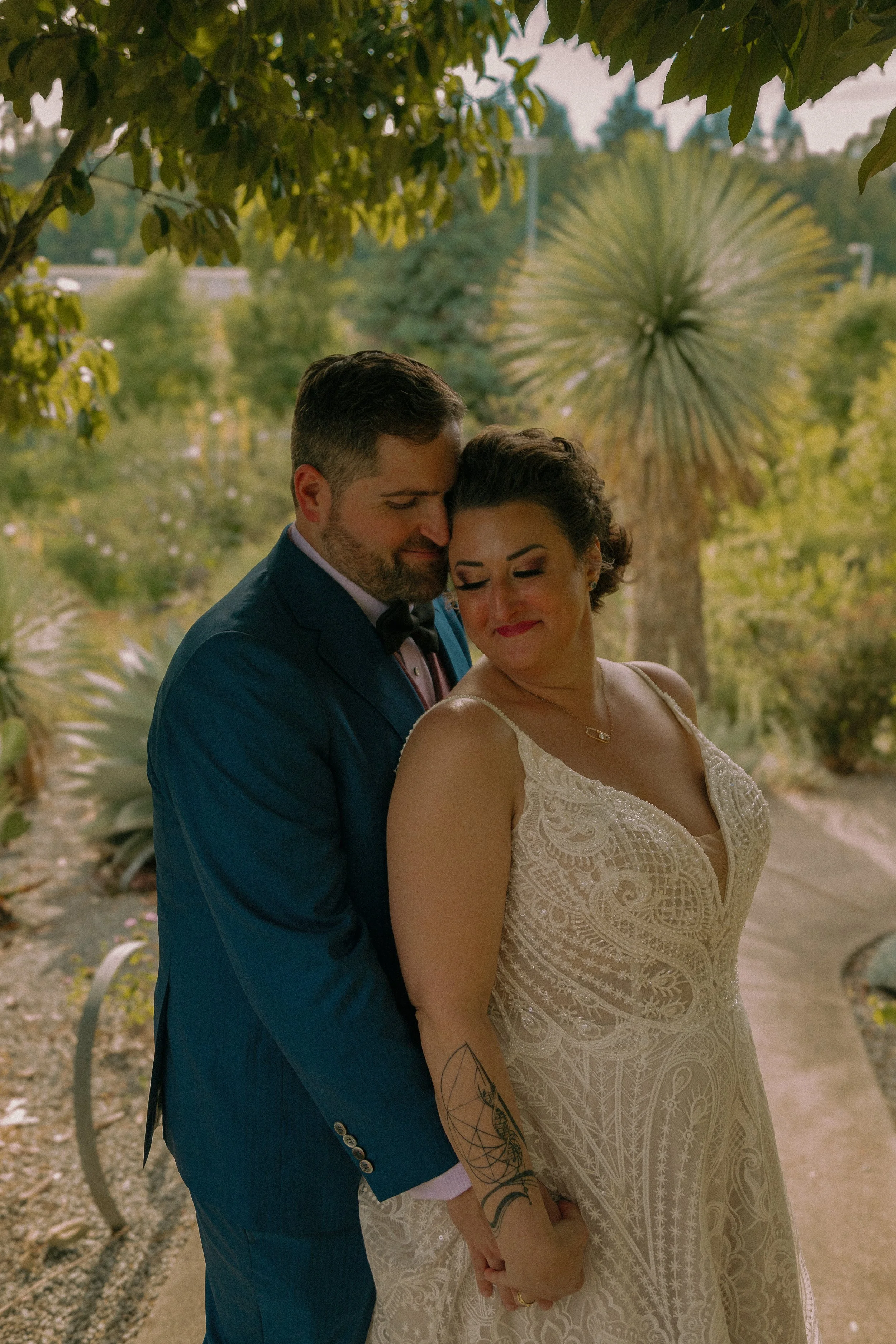 A couple embracing in a garden setting, with the woman wearing a lace wedding dress and the man in a dark blue suit.