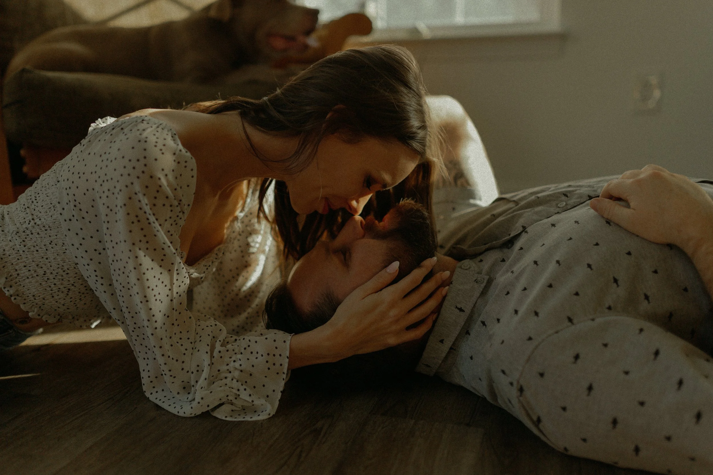 Couple lying on the floor, woman leaning over man with heads touching, in a cozy room with a dog on a couch in the background.