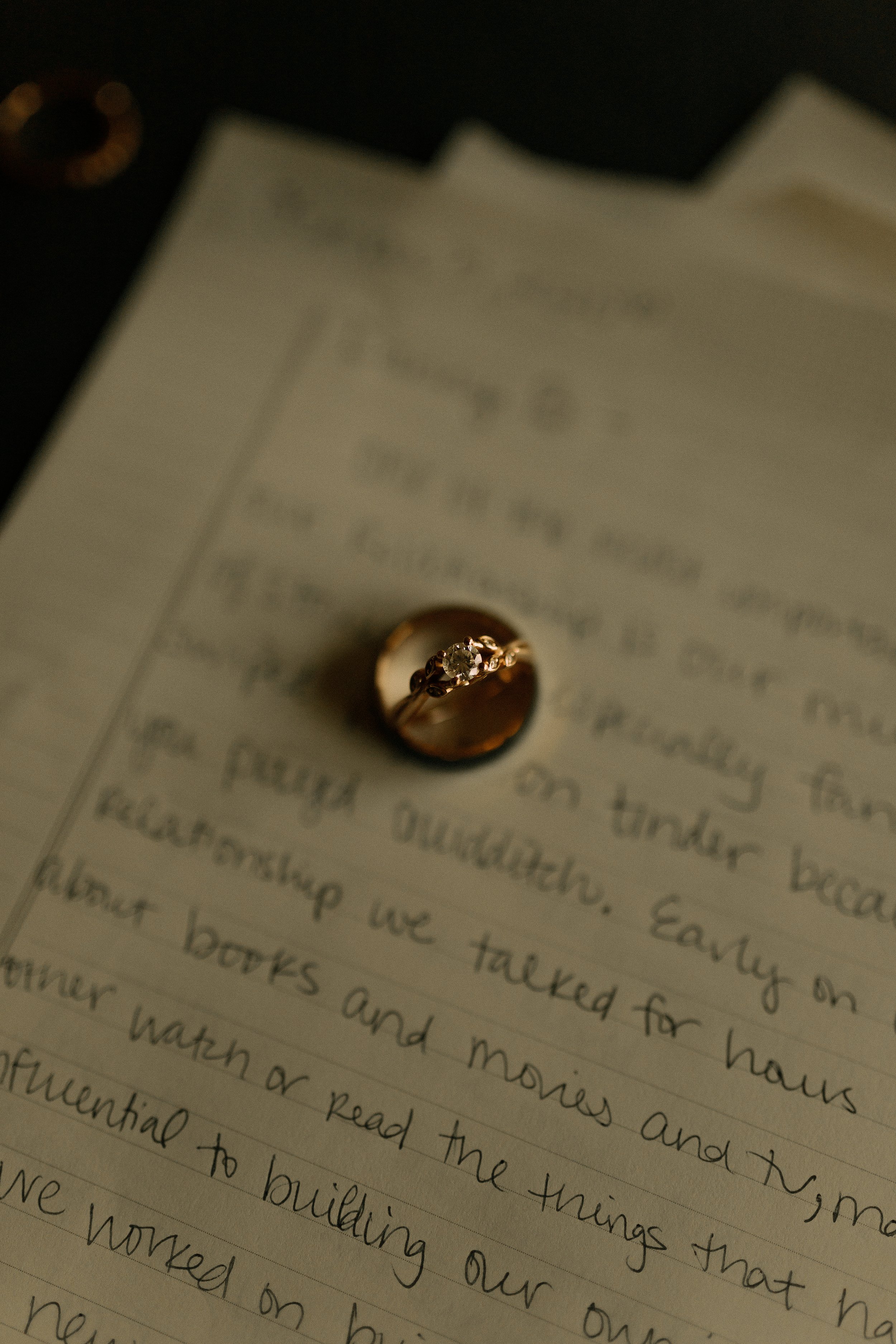 Close-up of a handwritten letter with an engagement ring placed on top, featuring a round diamond and decorative band. The letter is on lined paper.