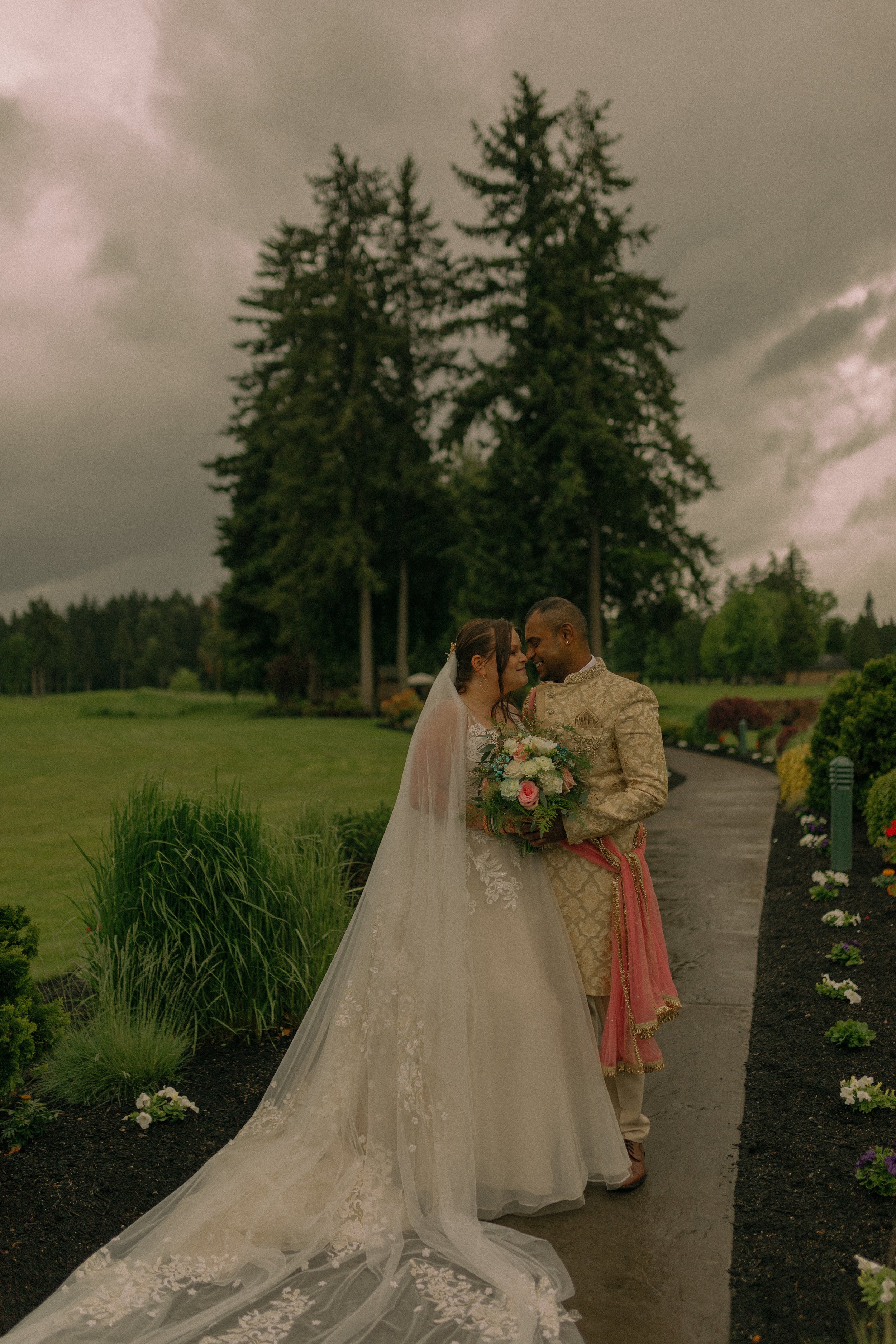 A bride and groom standing on a walkway in front of trees, with the bride wearing a long white wedding dress and holding a bouquet of flowers. The groom is in traditional attire with a pink sash. The setting is outdoors with greenery around.