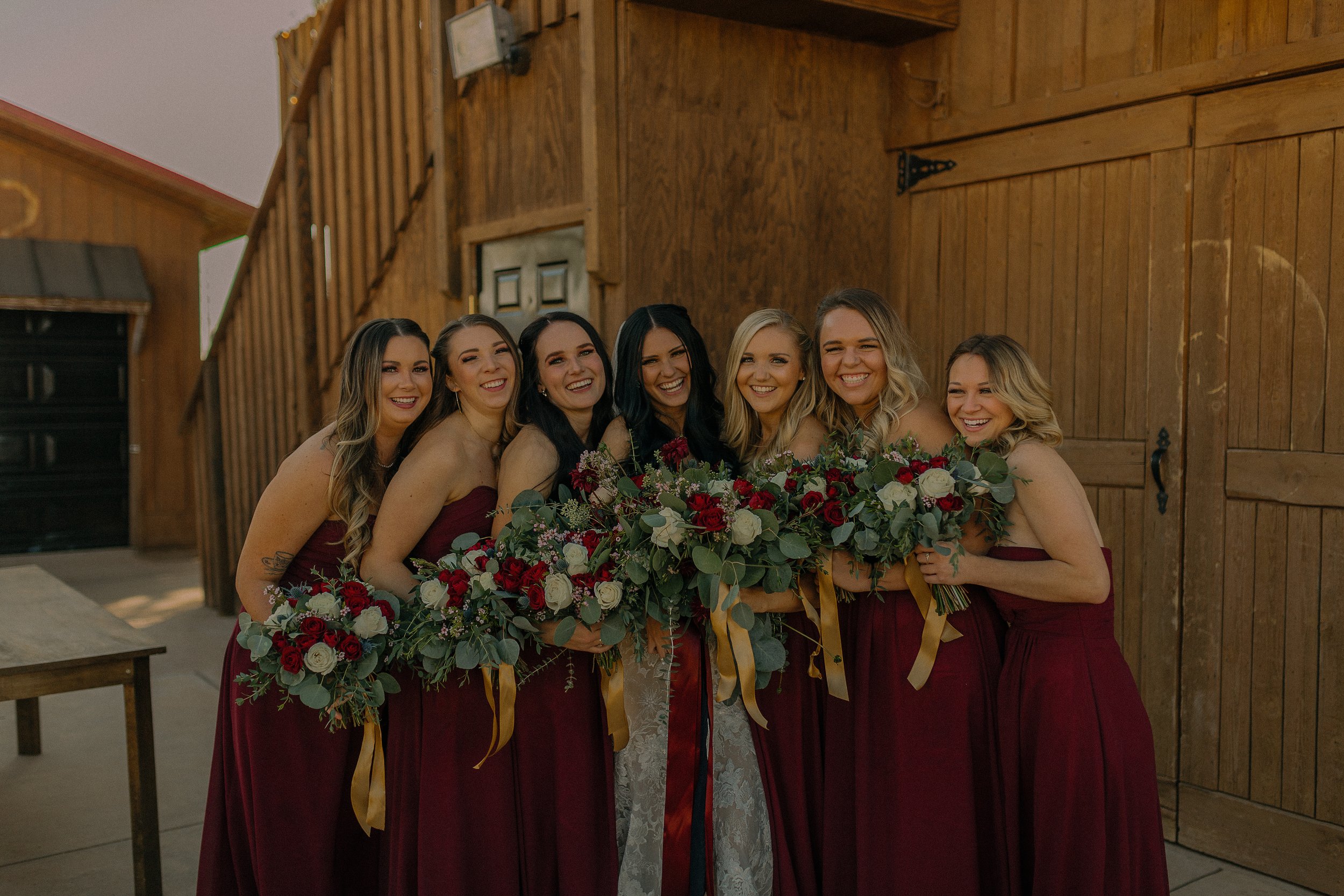 Bridesmaids in burgundy dresses holding bouquets, smiling next to a wooden barn.