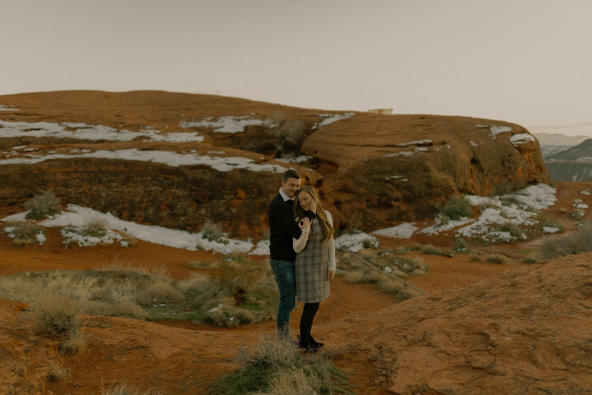Couple standing on a rocky terrain with patches of snow in a desert landscape.