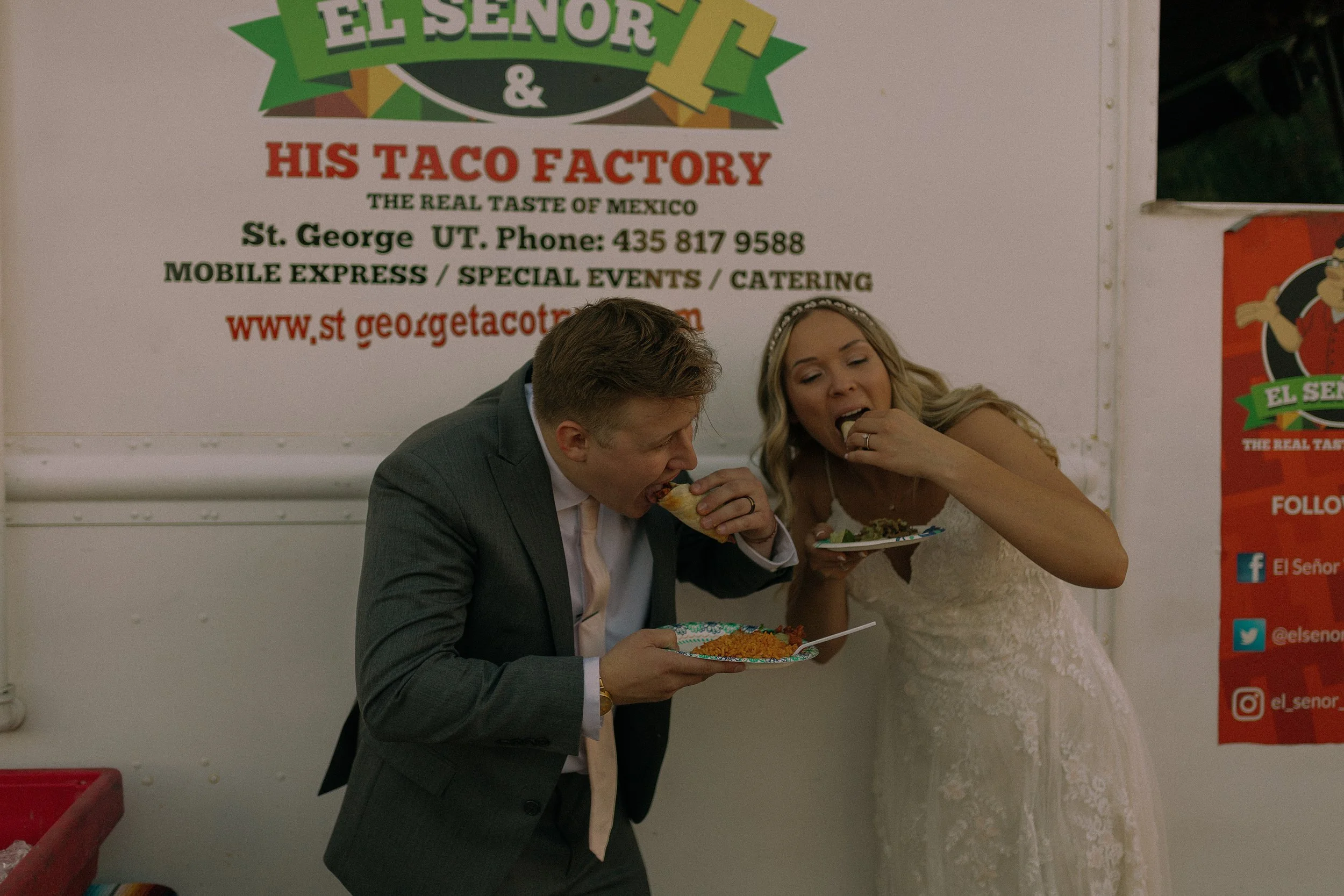A couple in wedding attire eating tacos from plates in front of a taco truck.
