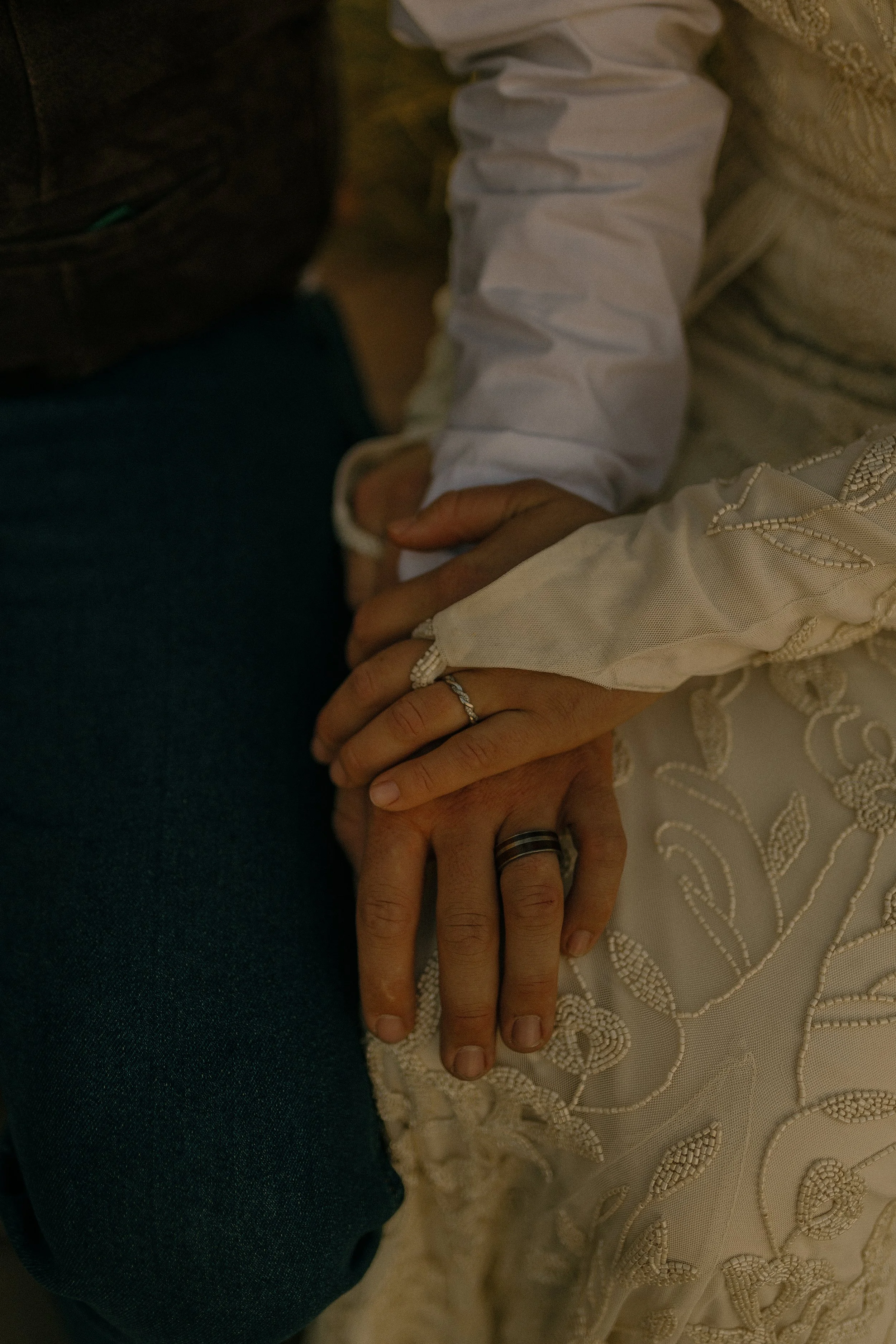 Couple's hands with wedding rings resting on a beaded dress and jeans