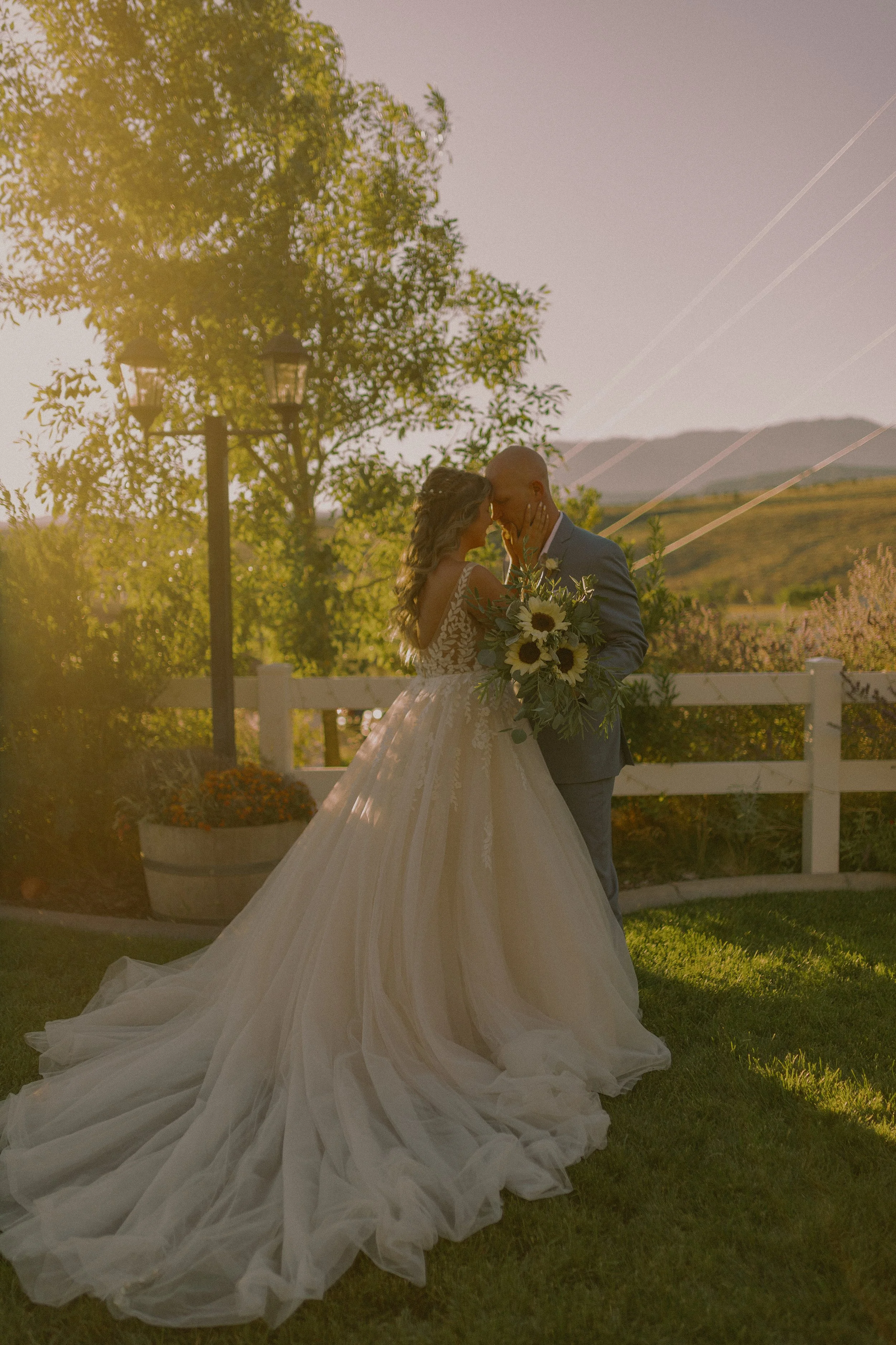 Bride and groom embracing in a garden at sunset, with the bride wearing a long, flowing wedding dress and holding a bouquet of sunflowers.