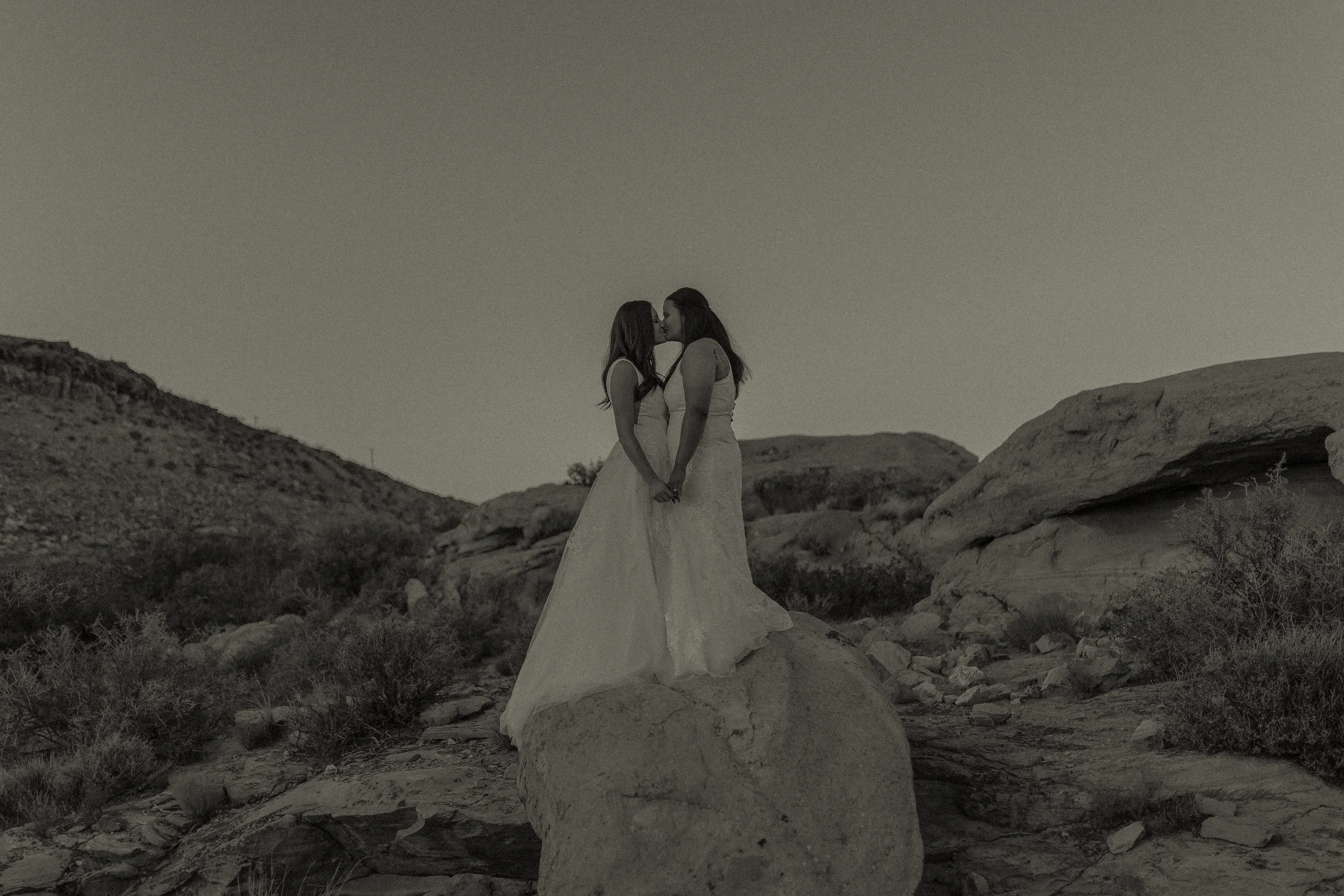 Two people in wedding dresses kissing on rocky terrain with a desert backdrop.