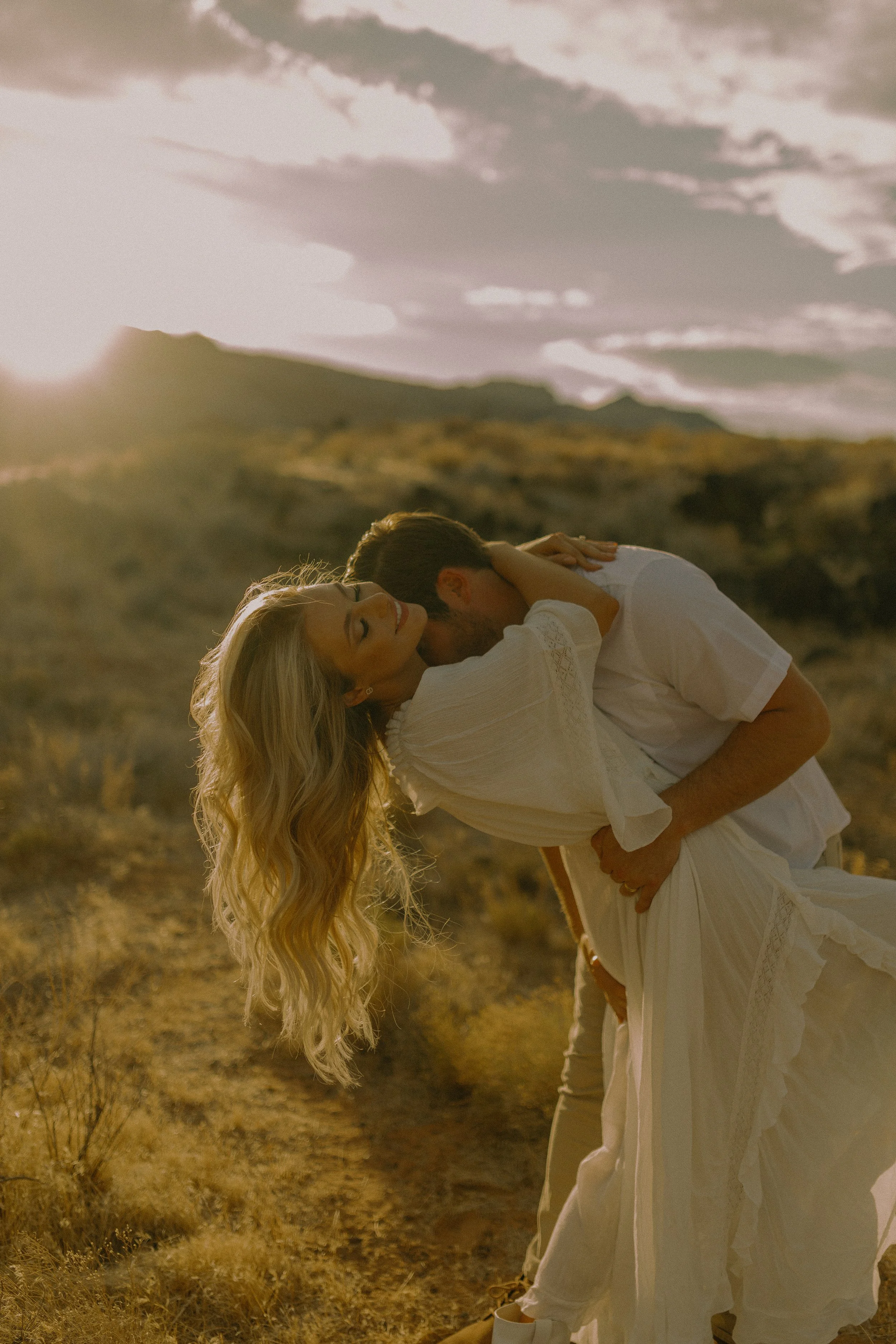 Couple embracing at sunset in a field, woman in white dress being dipped by man, warm glow.