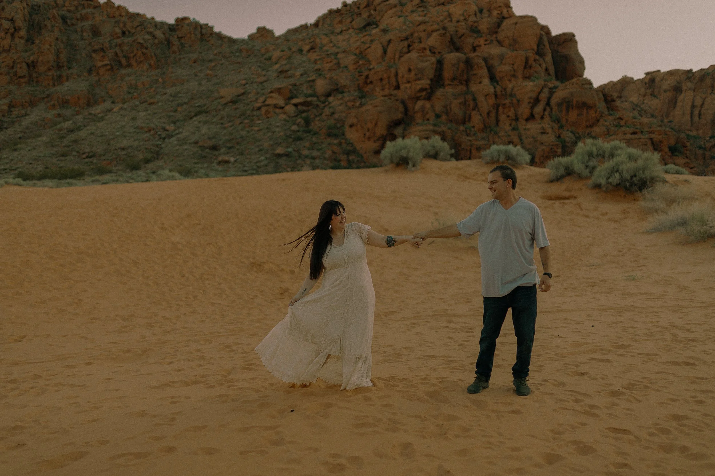 A man in a gray shirt and jeans holds hands with a woman in a white dress walking on a sandy desert landscape with rocky hills in the background.