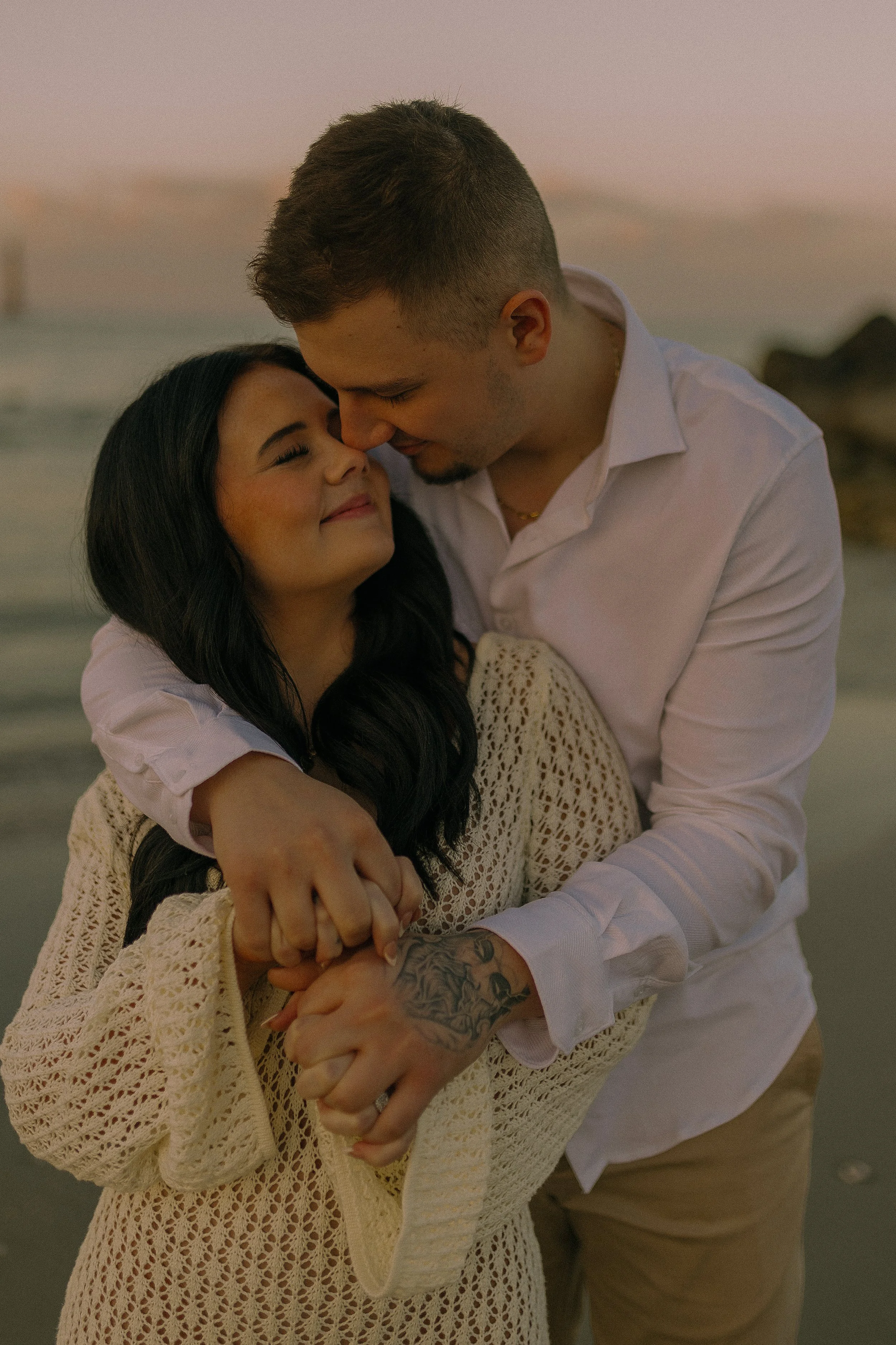 A couple embracing on a beach, with the man in a white shirt hugging the woman wearing a crochet sweater, during sunset.