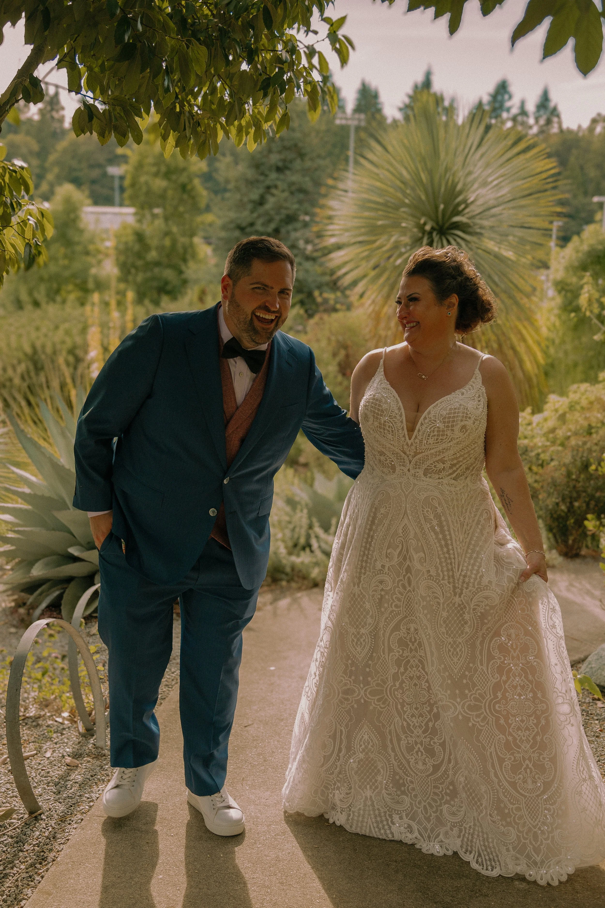 Bride and groom smiling in an outdoor setting, with greenery and trees in the background. The bride is wearing a lace wedding dress, and the groom is in a blue suit.