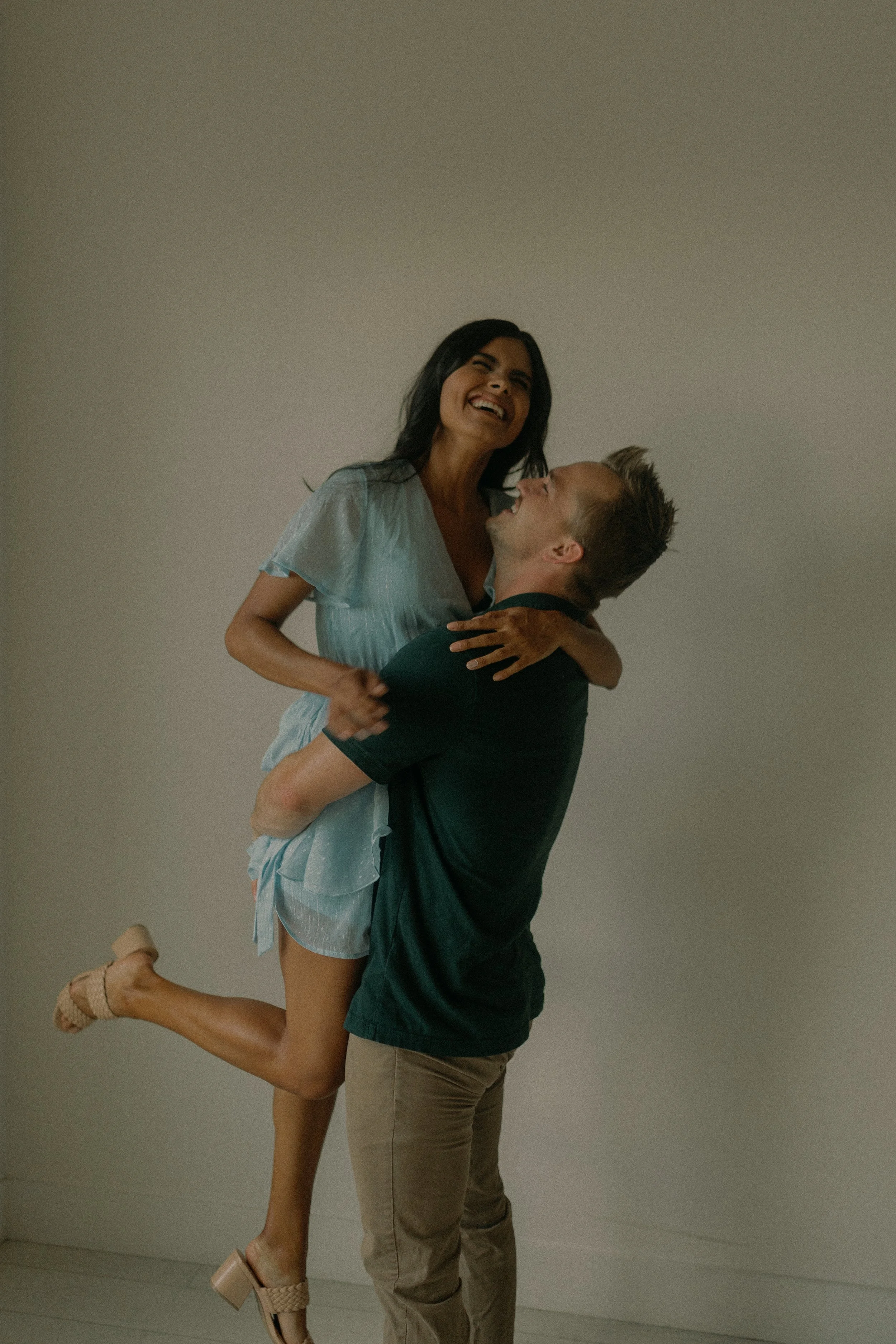 A woman in a light blue dress being lifted by a man in a dark green shirt, both smiling joyfully.