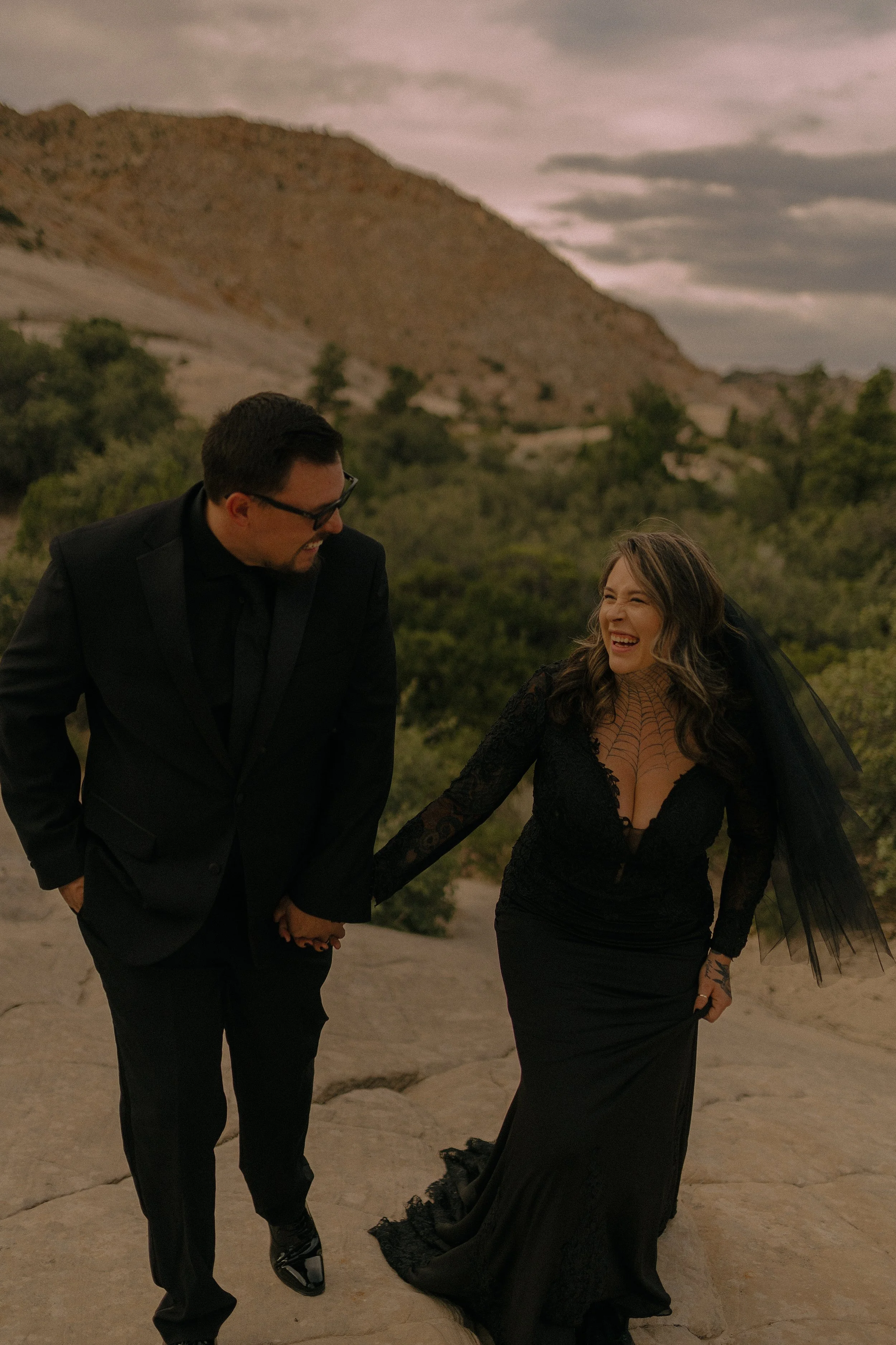 A couple wearing black formal attire is walking outdoors on rocky terrain with a mountainous backdrop. The woman is in a black dress with a lace design and veil, and the man is in a black suit and tie. Both appear joyful.