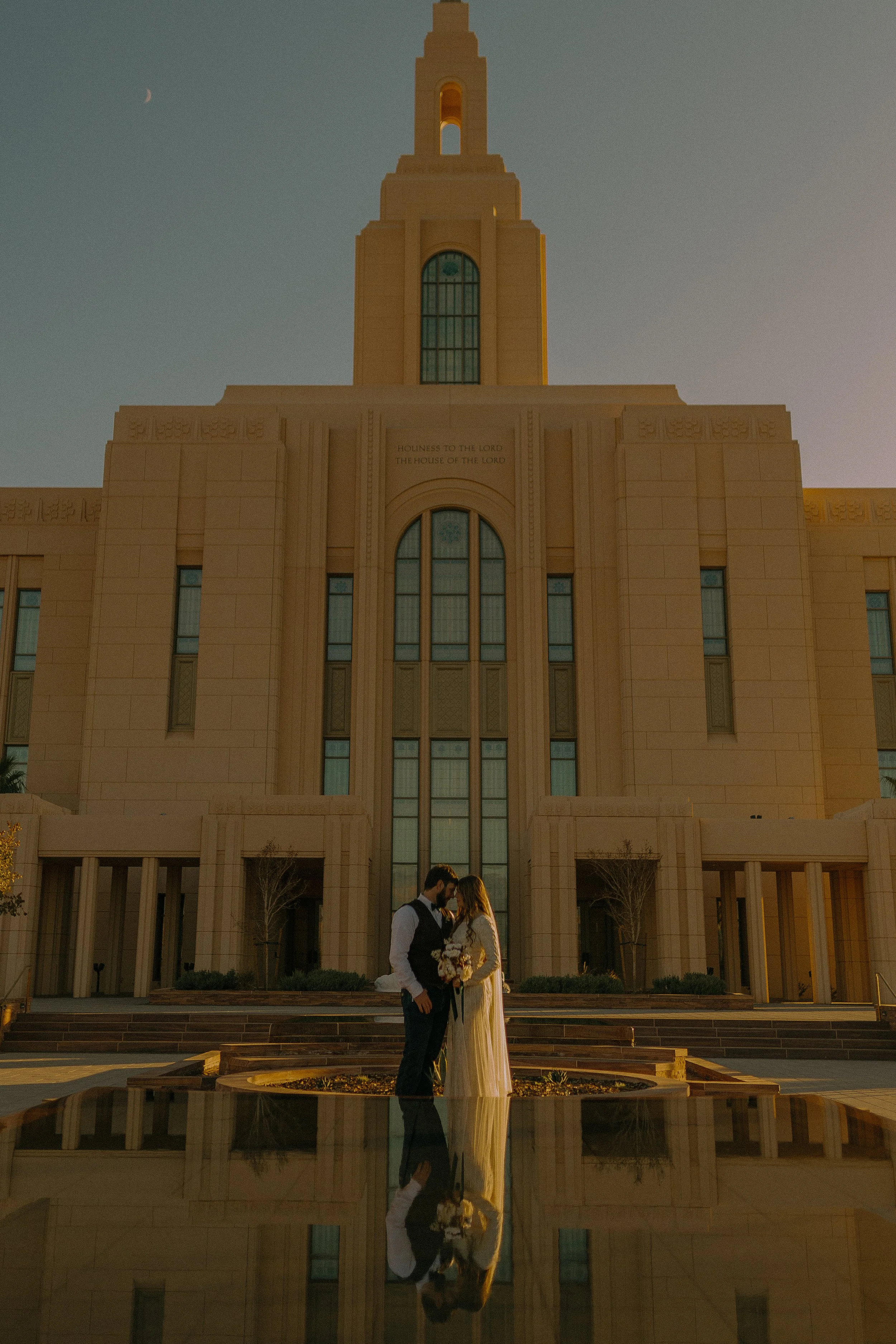 A couple in wedding attire embracing in front of a large, beige-colored temple at sunset, with a clear reflection in a pool.