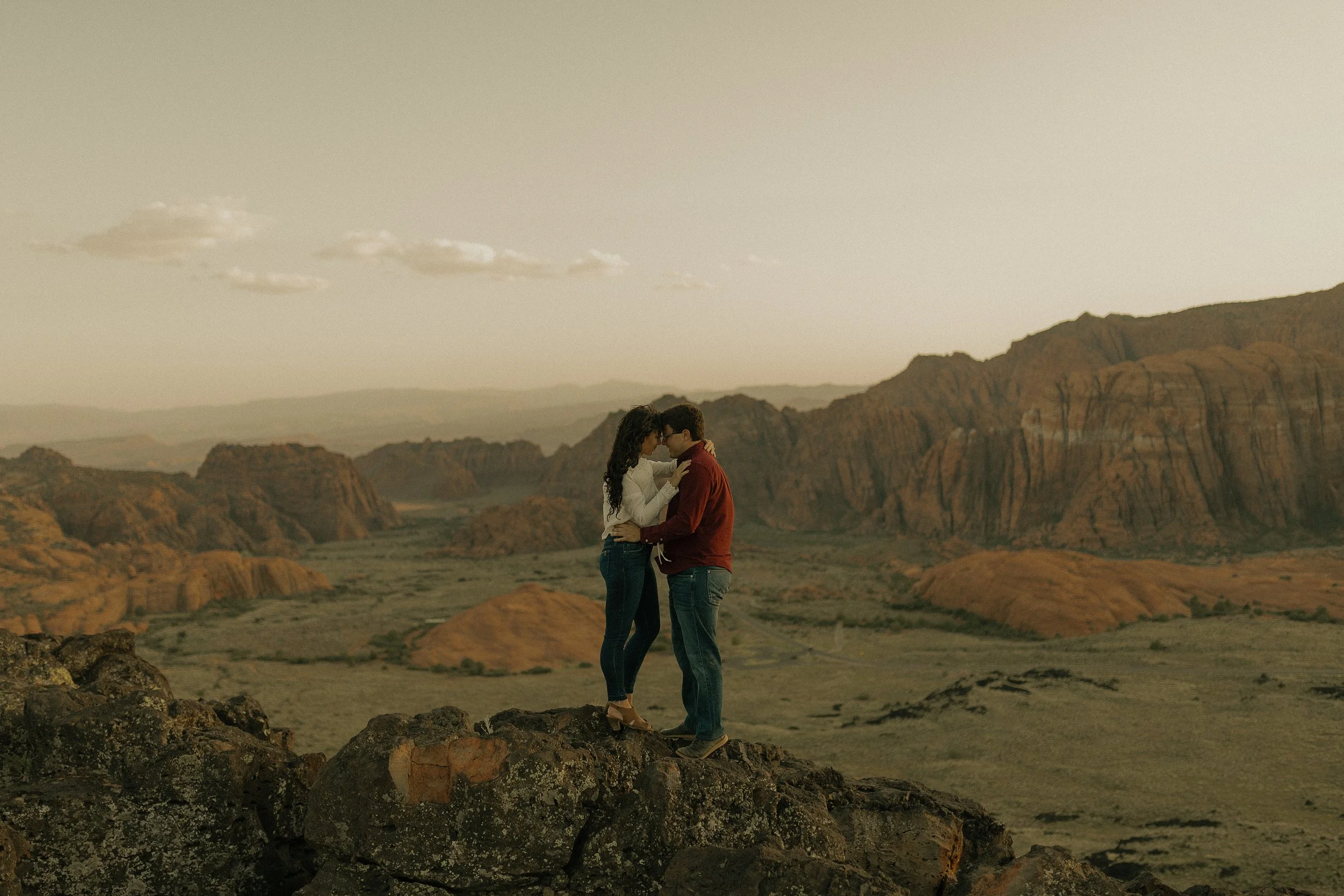 Couple embracing on rocky cliff with desert landscape and mountains in the background.