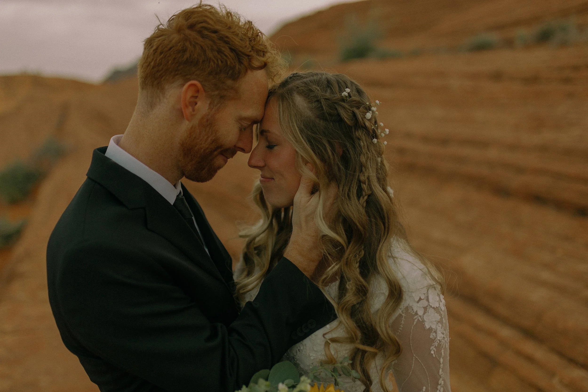 A groom in a black suit and a bride in a white lace dress embracing closely with their foreheads touching, set against a desert landscape.