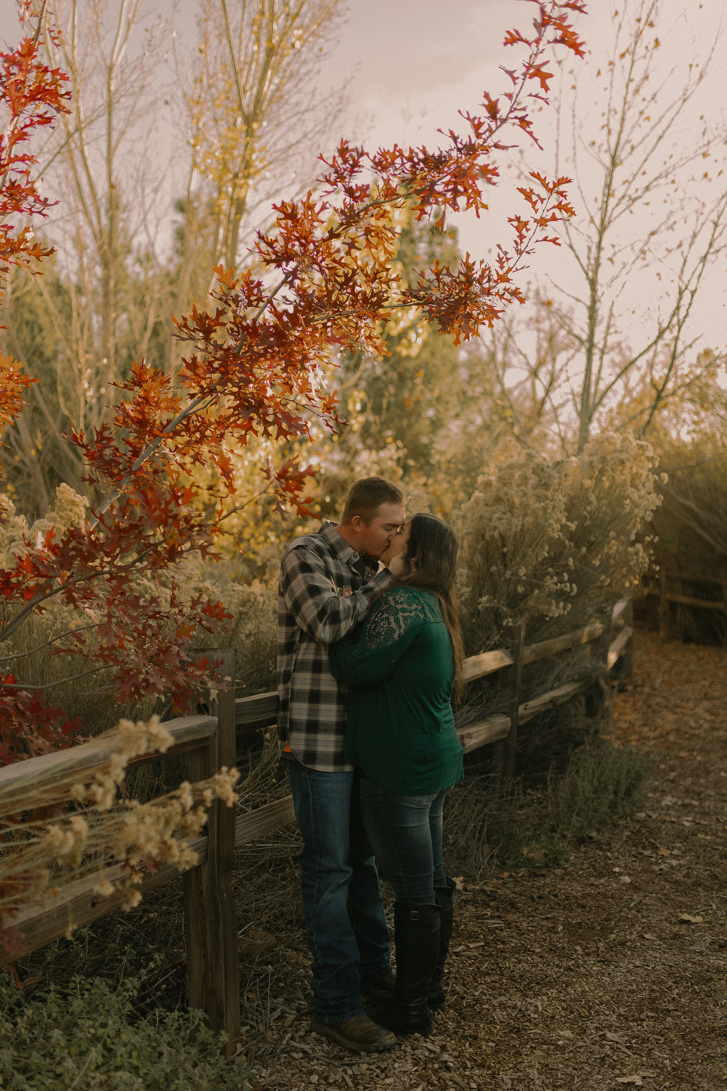 A couple kissing in a rustic outdoor setting with fall foliage.