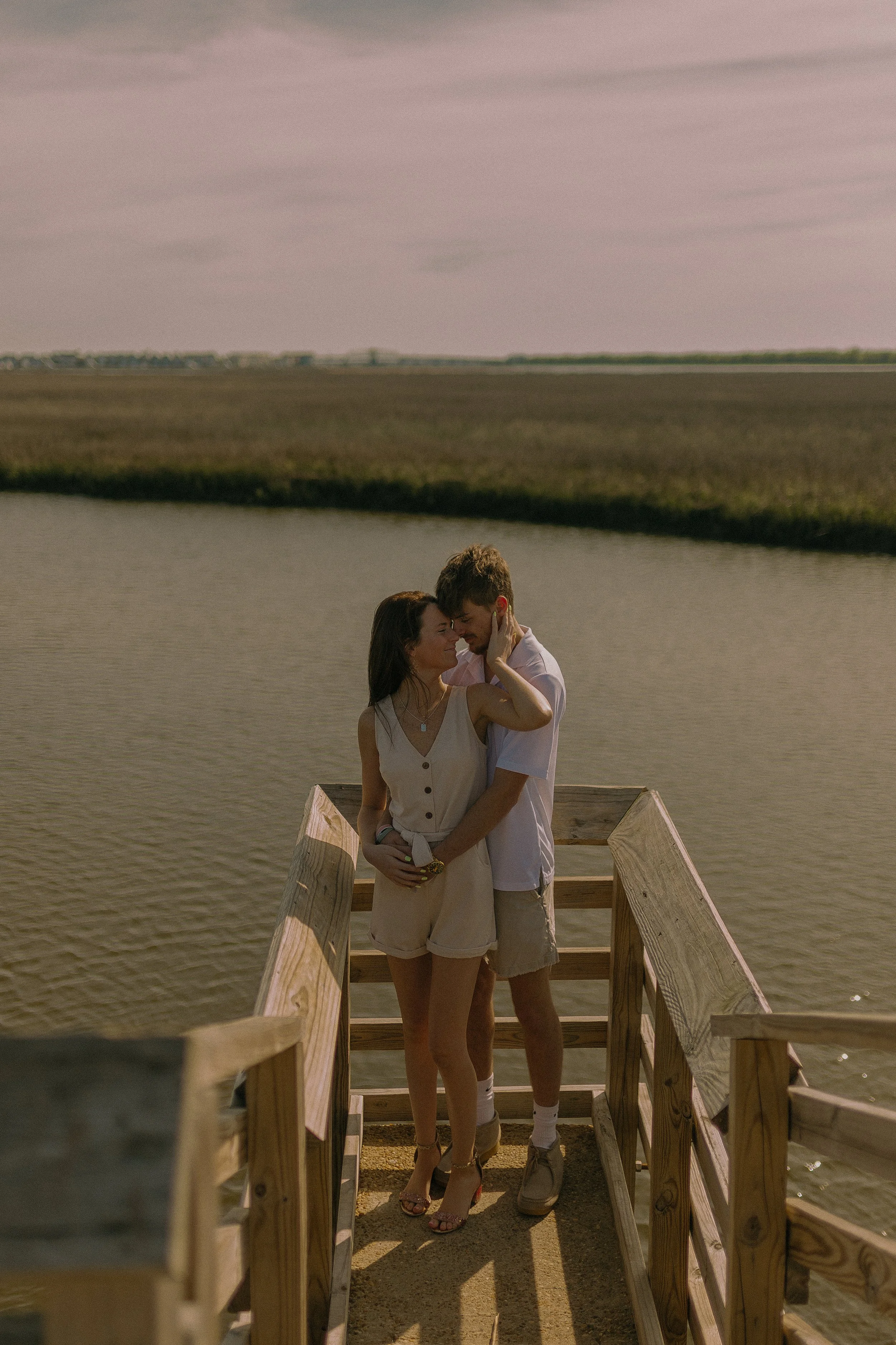 Couple embracing on a wooden boardwalk by a waterway under a cloudy sky.