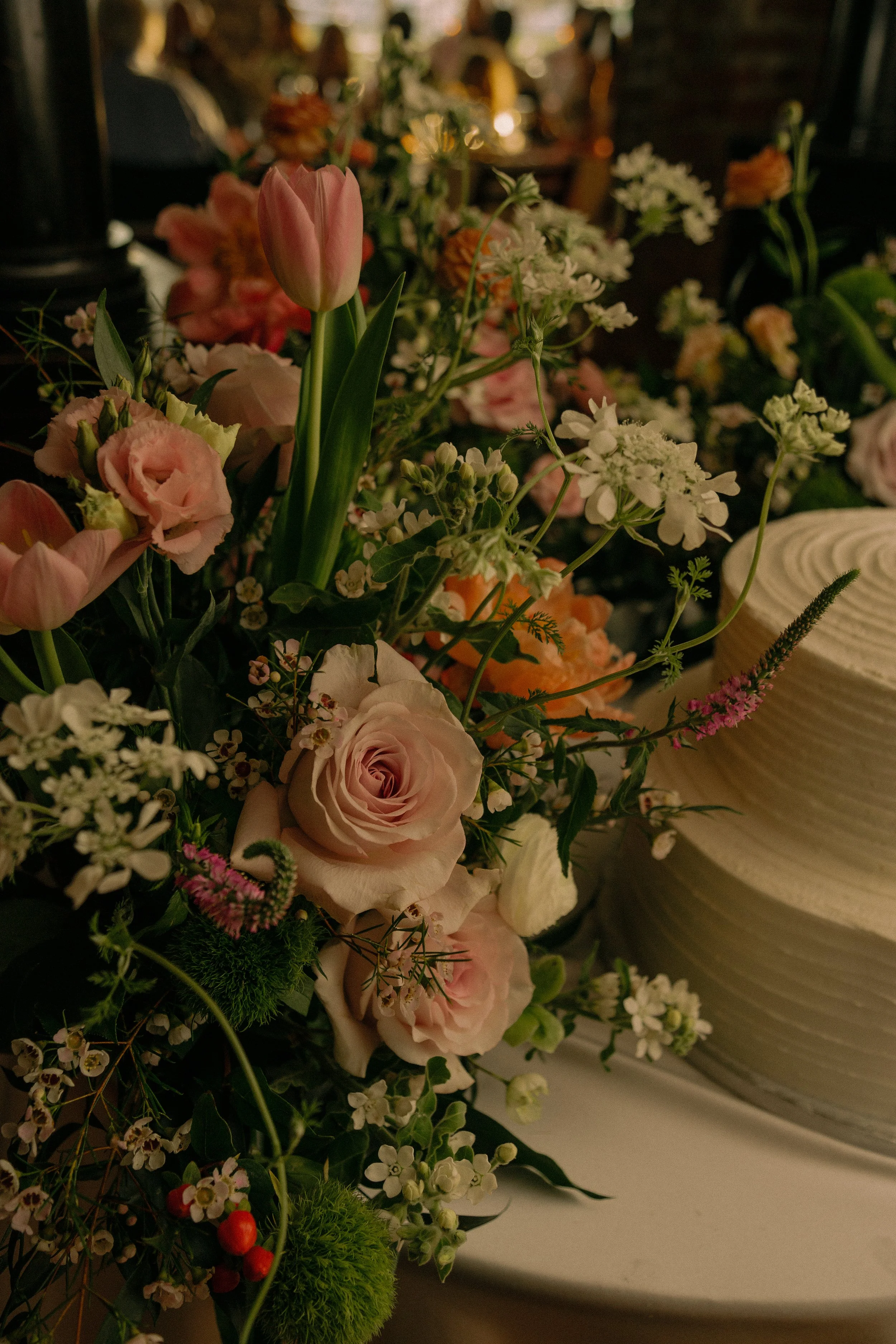 Bouquet of pink roses, tulips, and various wildflowers next to a white tiered cake.