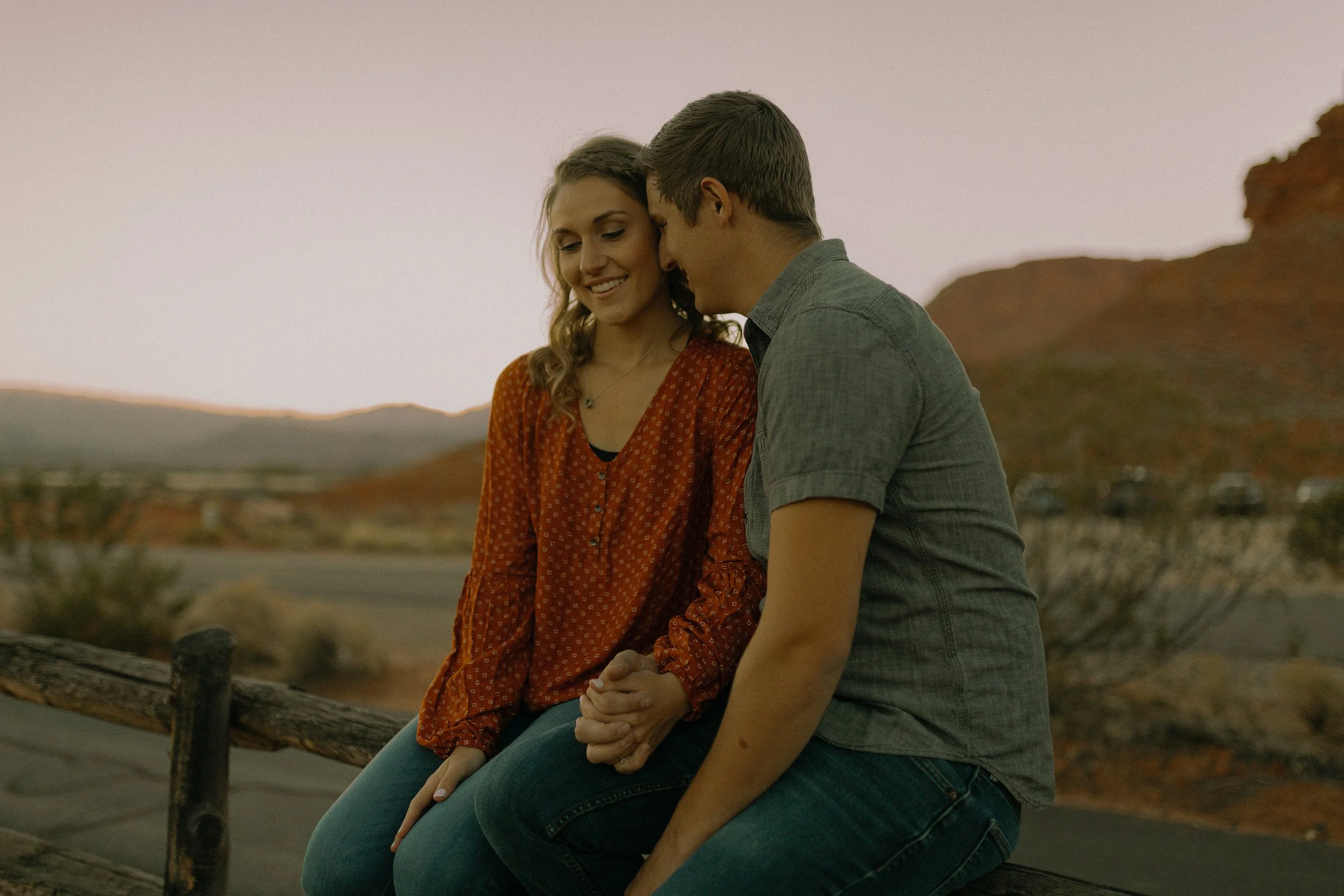 A couple sitting closely on a wooden fence in an outdoor setting, holding hands and appearing affectionate. The background features a scenic landscape with hills or mountains and a hazy sky.