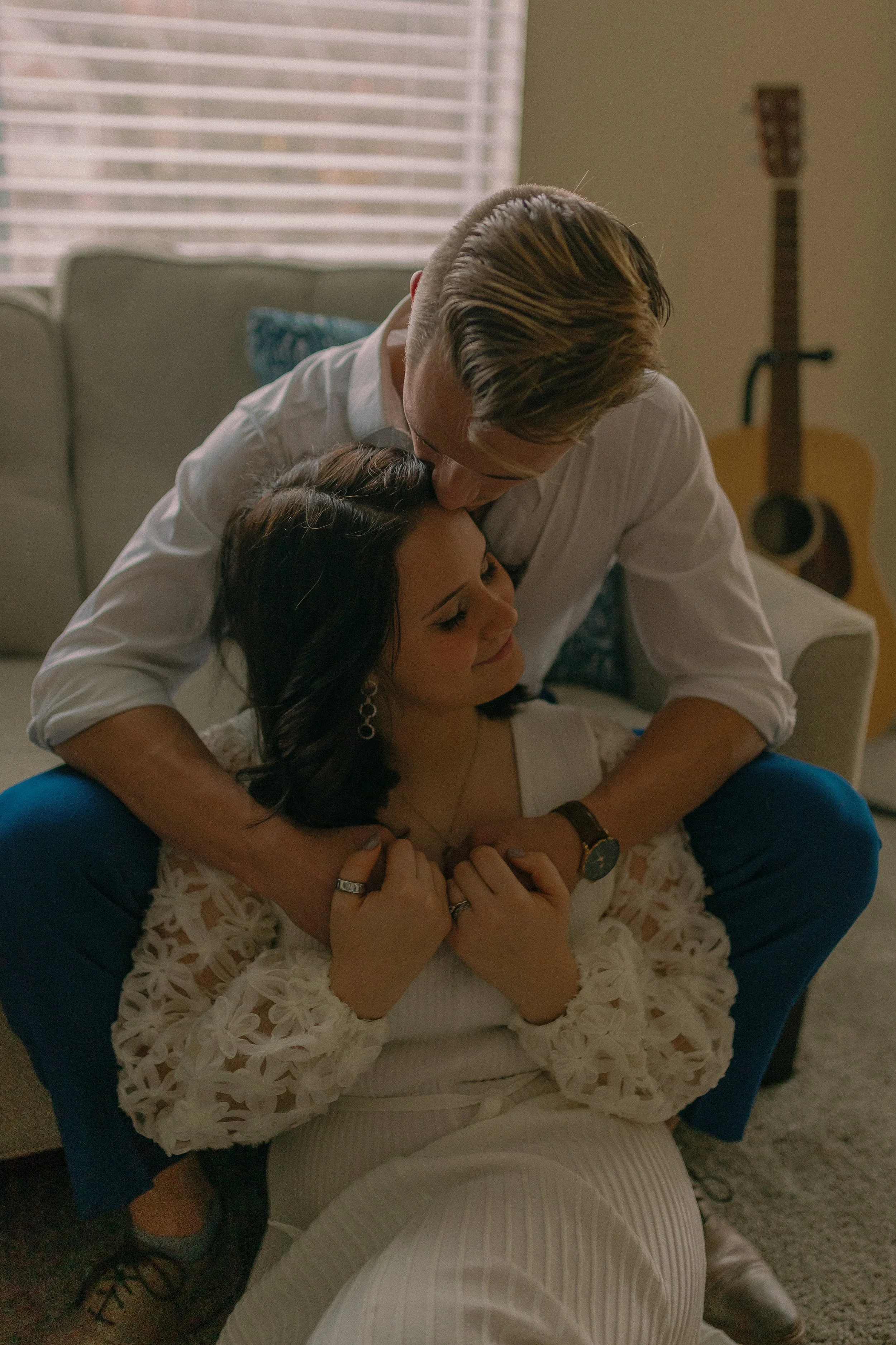 Couple sitting on a carpeted floor, man hugging woman from behind. Light from window blinds and acoustic guitar in background.